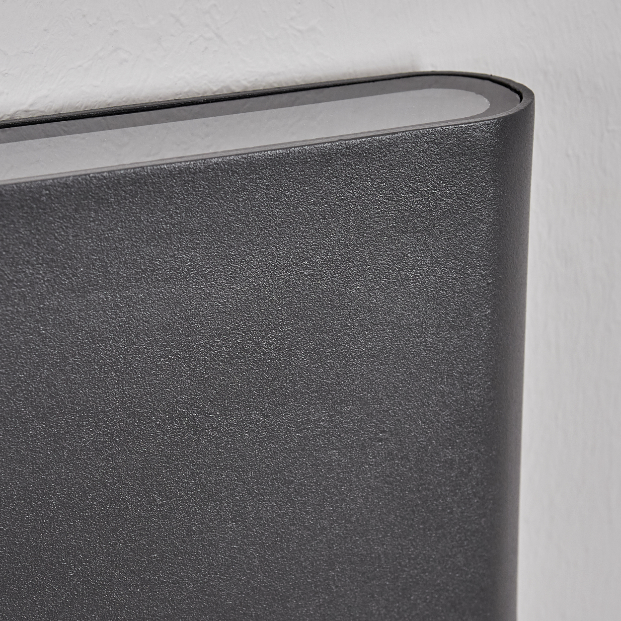 Dunkelgrauer, strukturierter Zylinder mit schwarzem Rand, an weißer Wand.