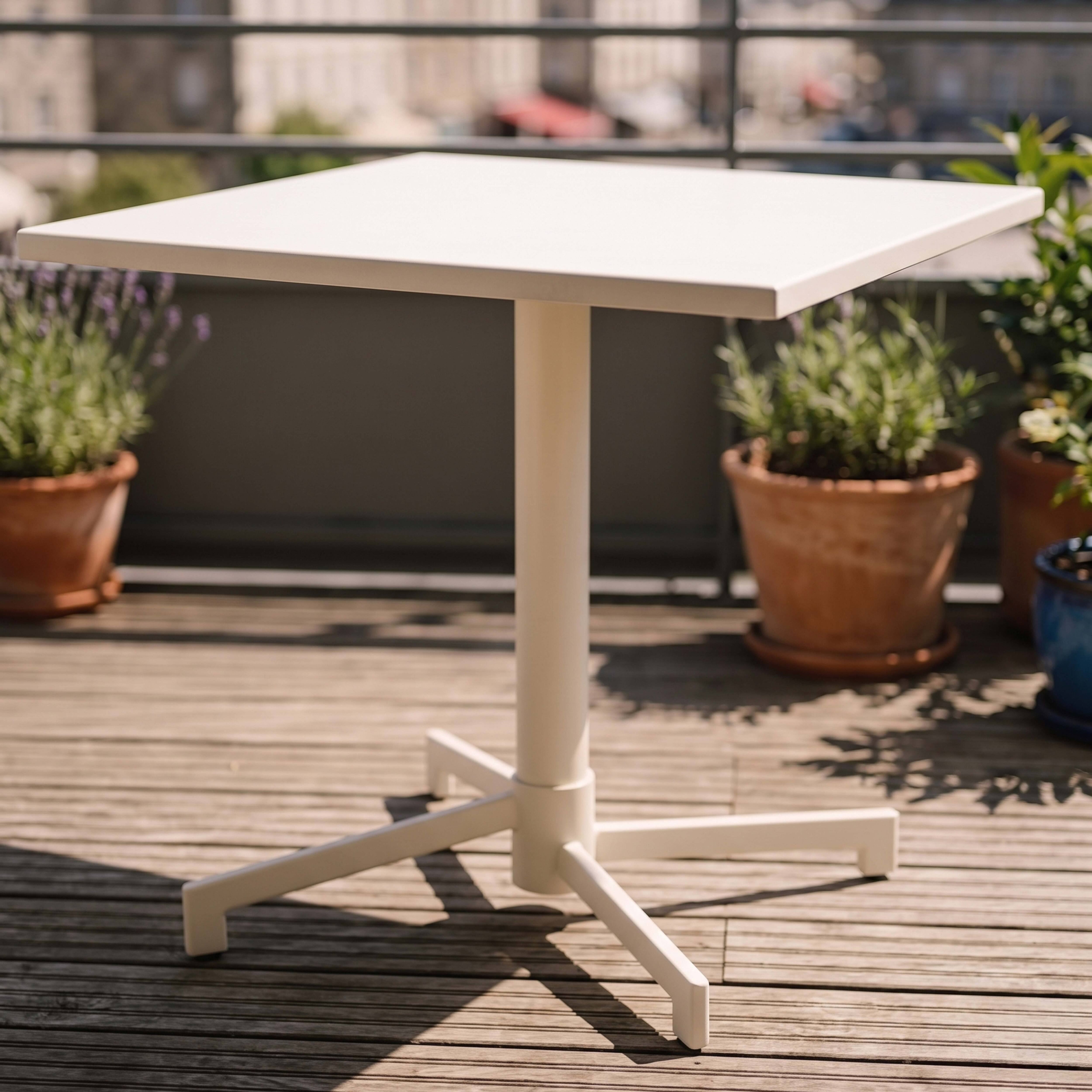 Ein weißer Tisch mit Kreuzfuß auf einer Holzterrasse, umgeben von Pflanzen.