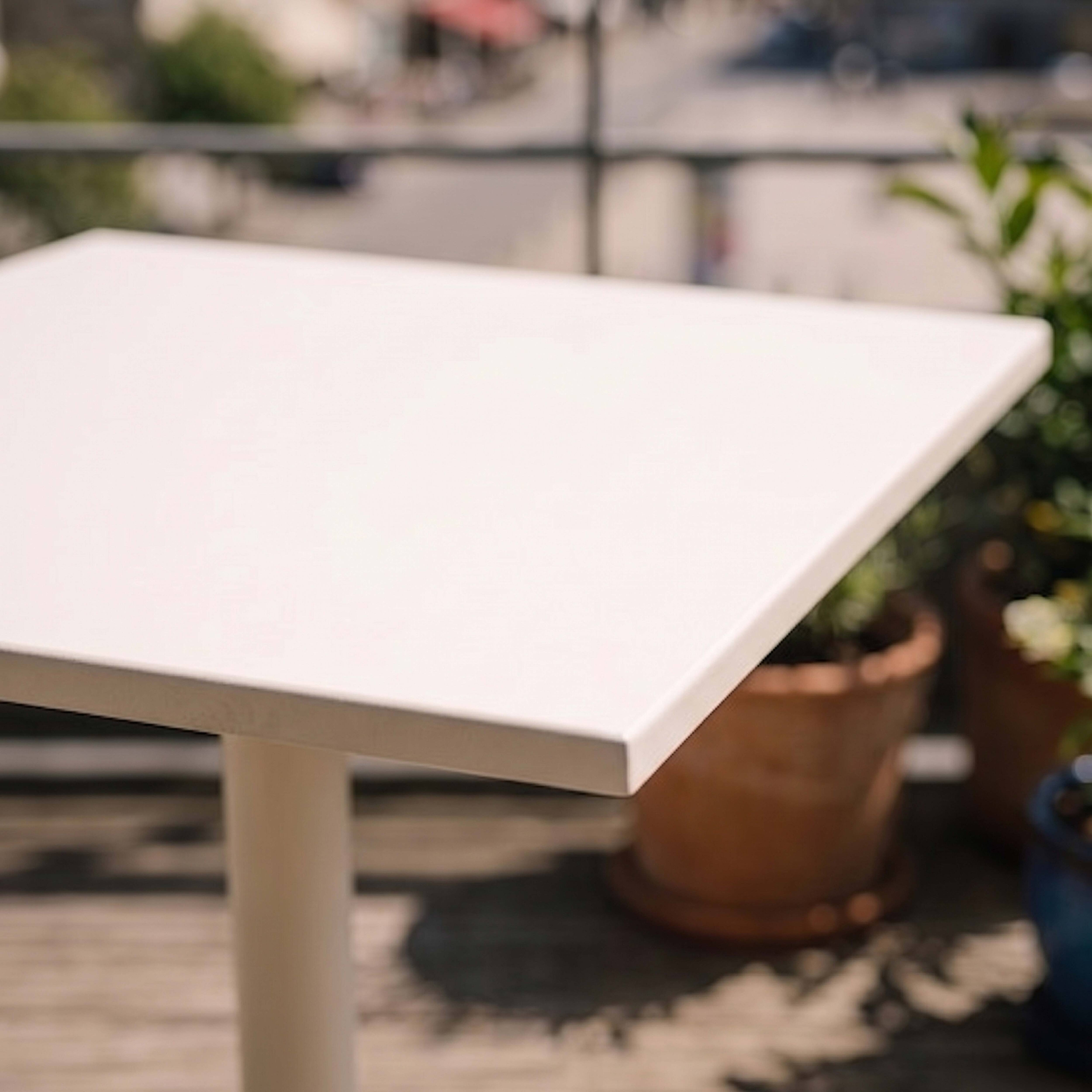 Ein weißer Tisch steht auf einer Holzterrasse, mit Pflanzen im Hintergrund.