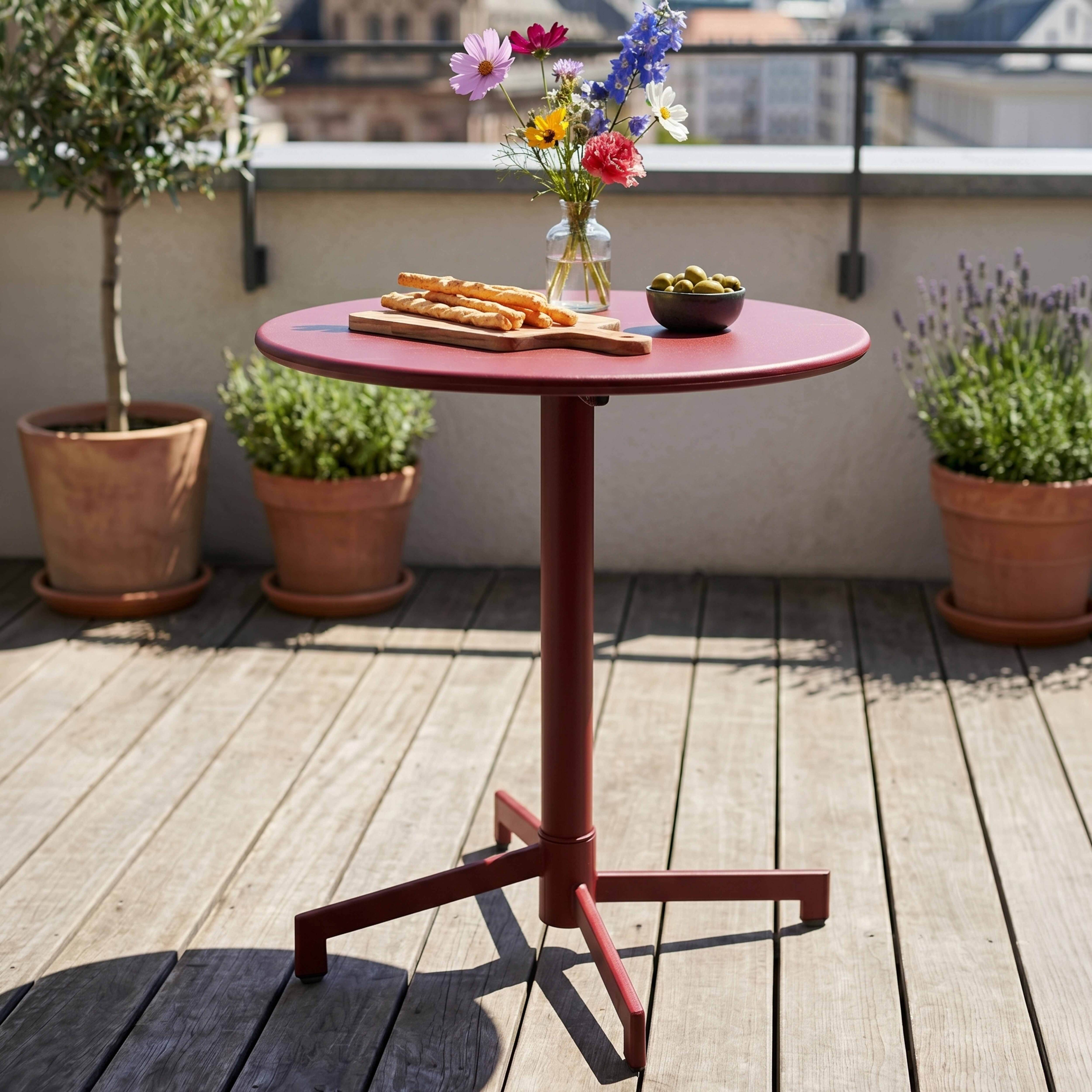 Roter Tisch auf Holzdeck mit Blumen, Brotstangen und Oliven. Pflanzen im Hintergrund.