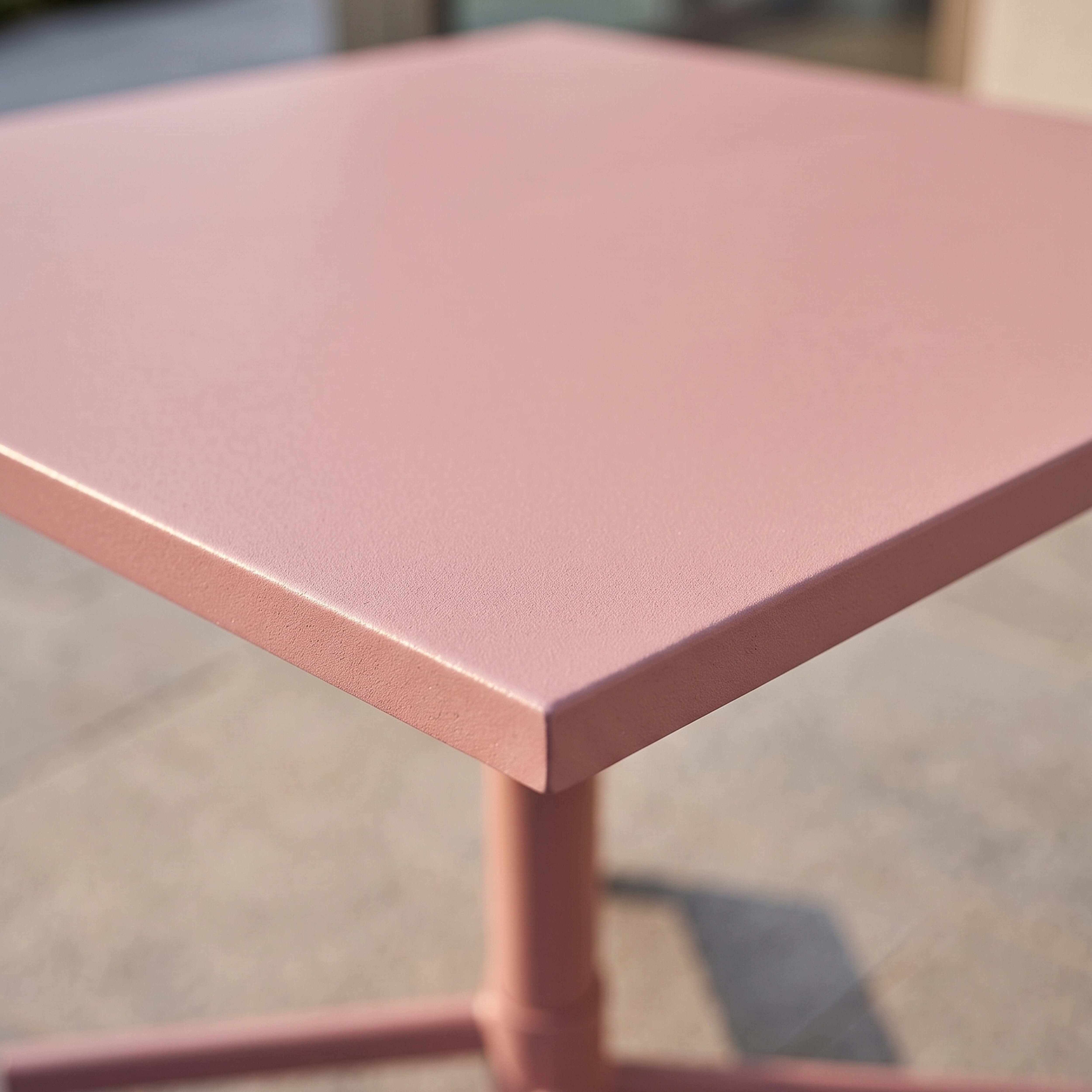 Ein rosafarbener quadratischer Tisch mit Metallbeinen auf einem gepflasterten Außenbereich.