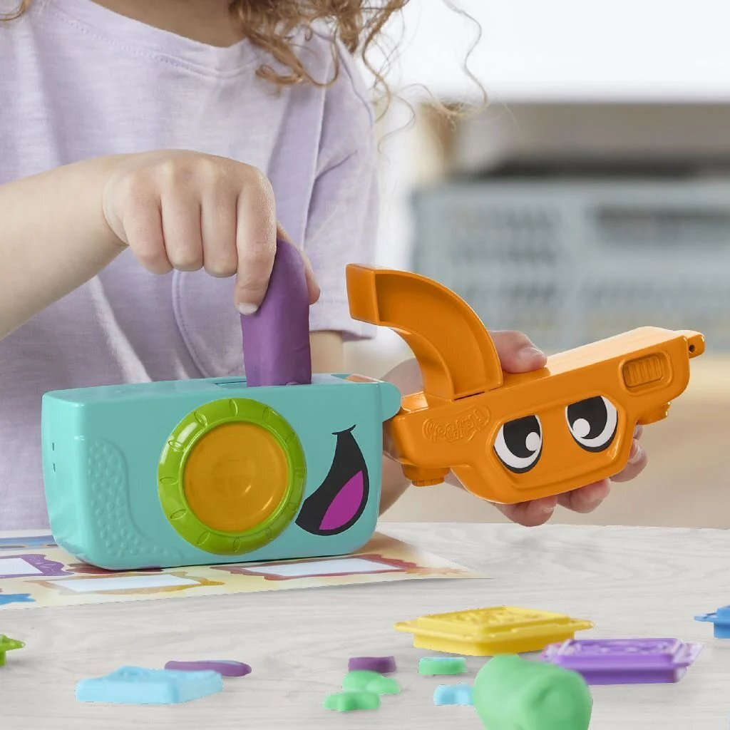 Un niño inserta plastilina morada en una cámara de juguete turquesa con cara feliz.