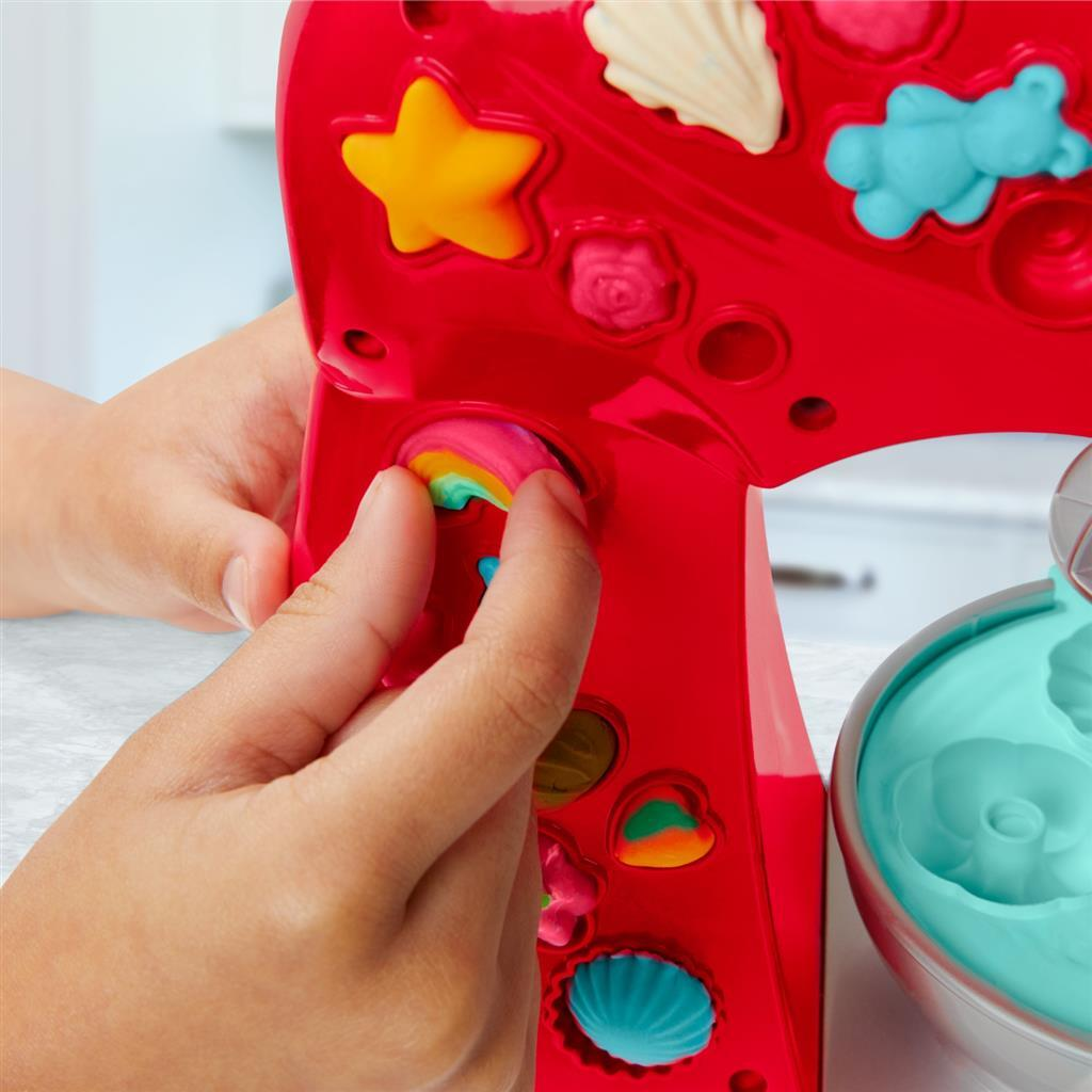 Kinderhände stecken ein regenbogenfarbenes Tonstück in einen roten Spielzeugmixer mit Formen.