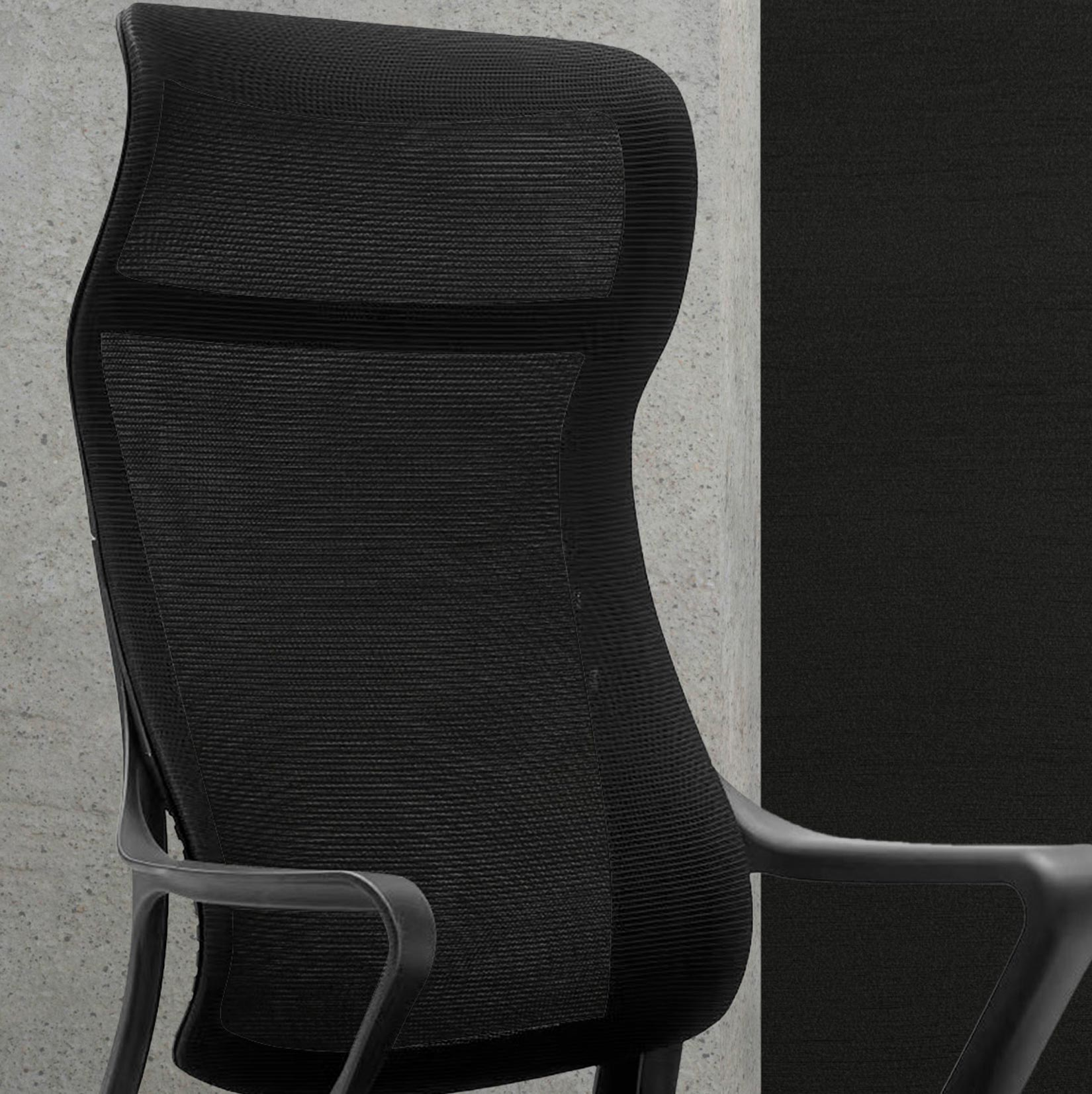 Silla de oficina negra con respaldo de malla y reposabrazos, contra muro de hormigón.