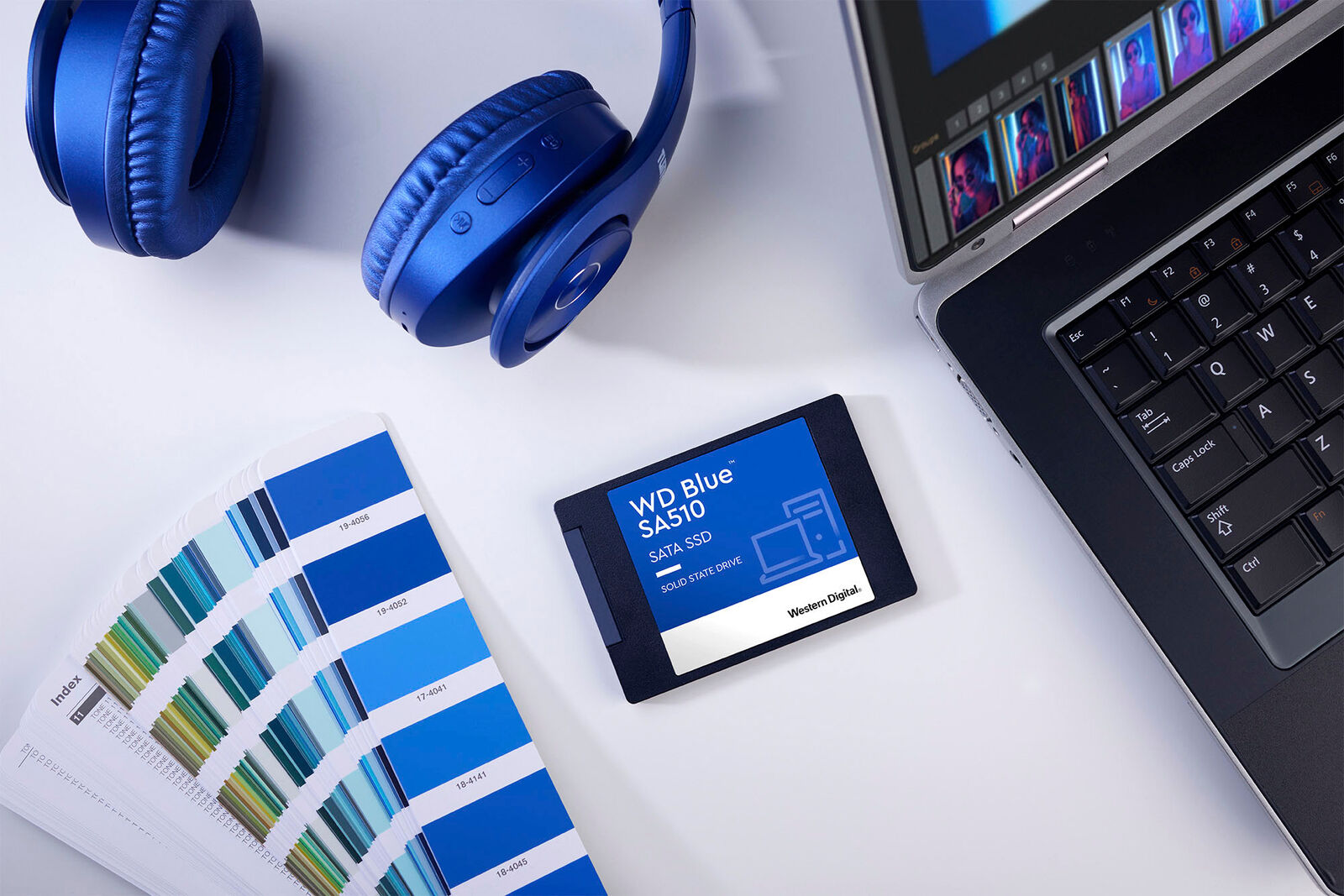 Cuffie blu, SSD WD Blue, campionario colori e laptop su scrivania bianca.