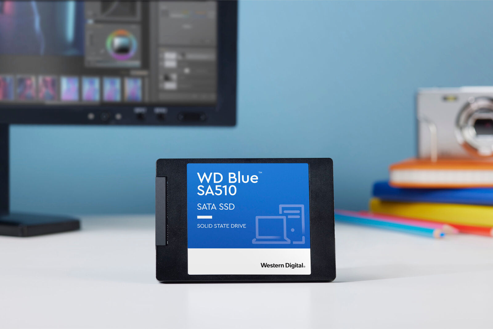 SSD SATA WD Blue SA510 su una scrivania con monitor del computer e fotocamera sullo sfondo.