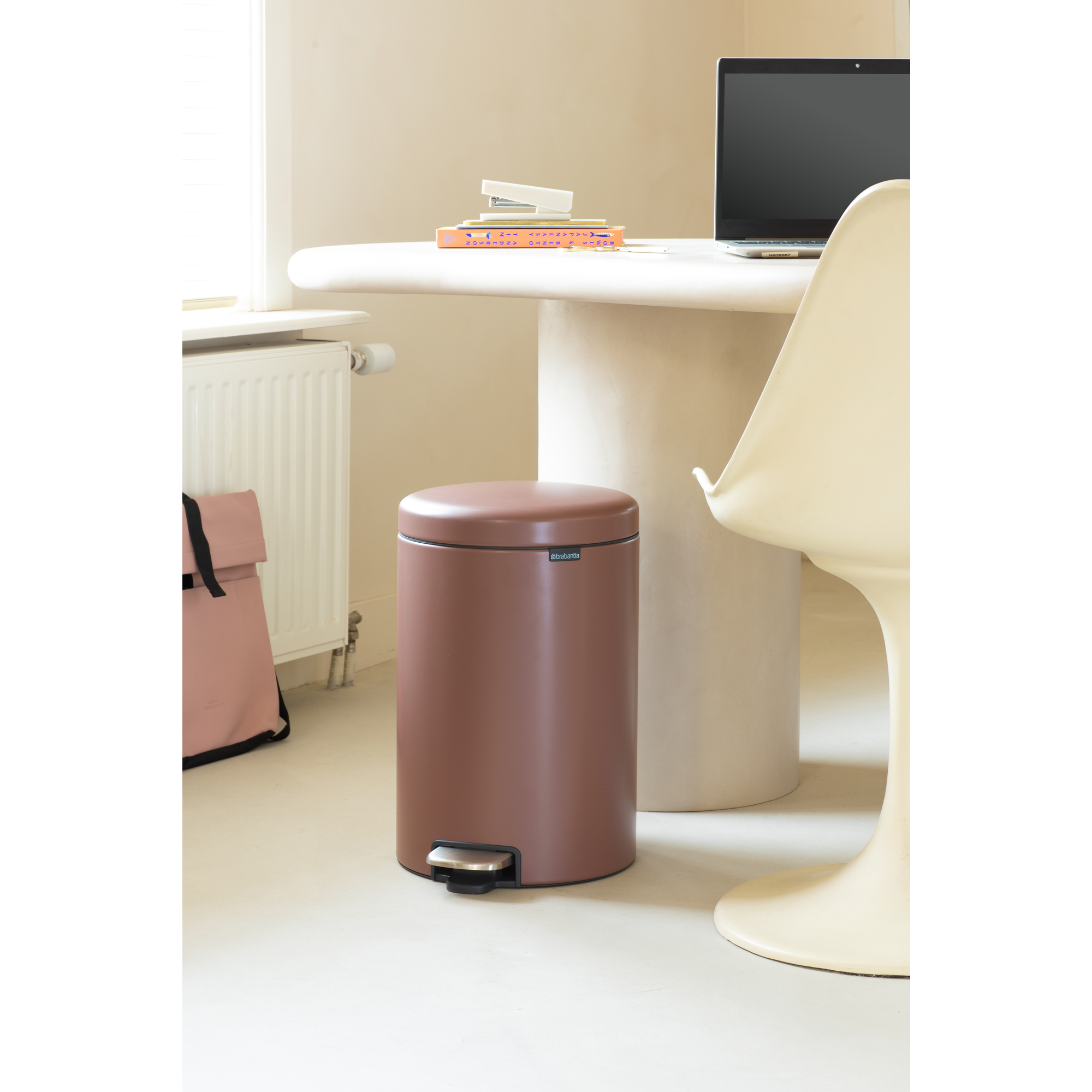 Een bruine prullenbak naast een radiator en een bureau met laptop.