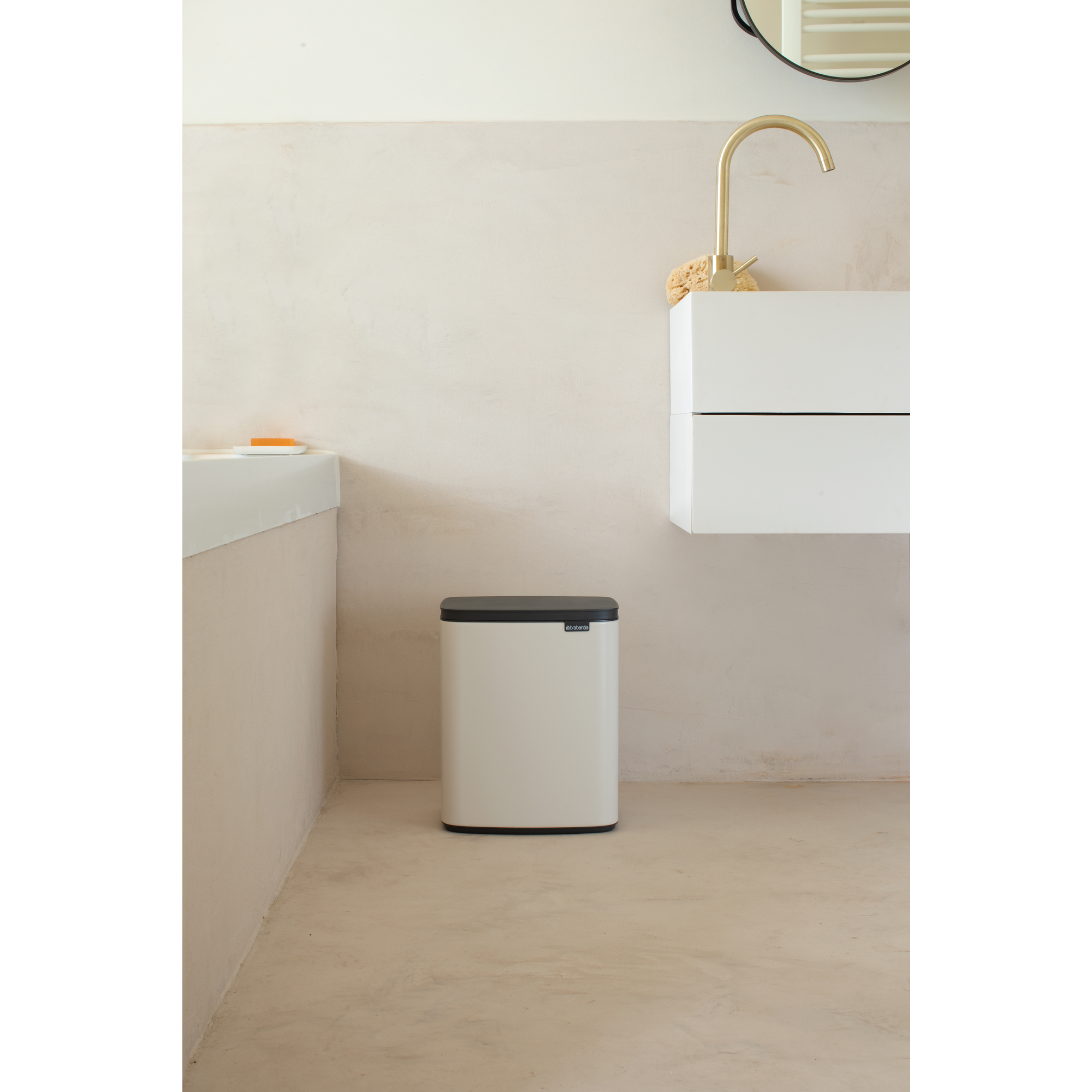 Een witte prullenbak met zwart deksel staat op een beige vloer naast een wastafelmeubel met messing kraan.