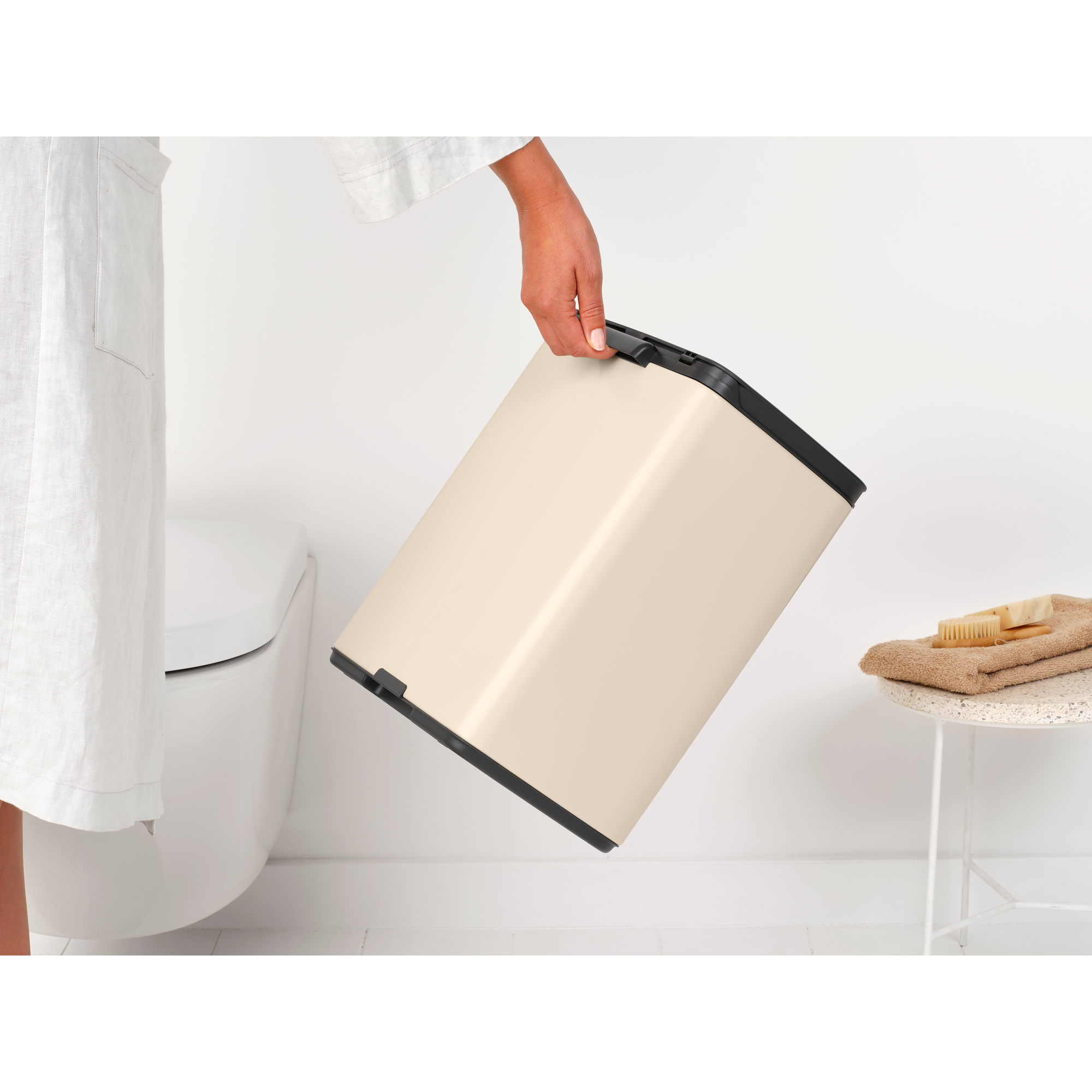 Een hand houdt een crèmekleurige prullenbak naast een toilet vast.