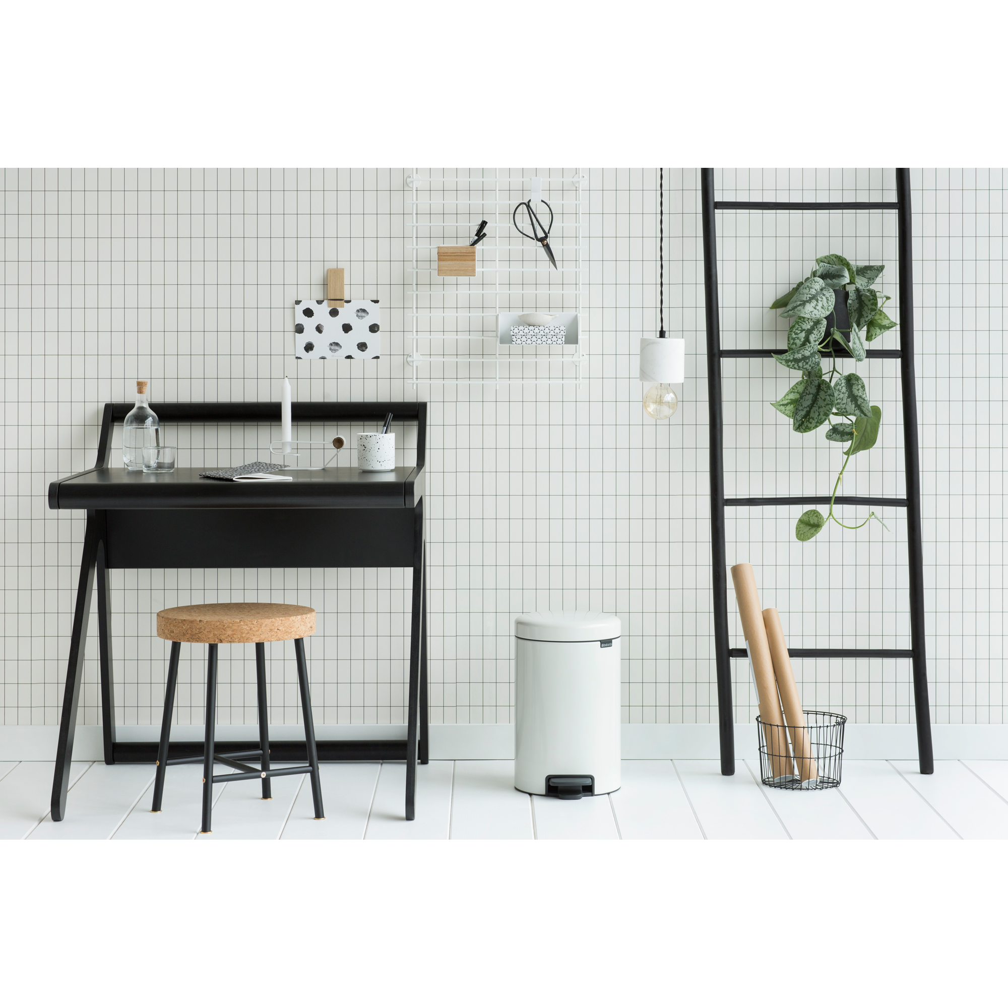 Een zwart bureau met een kurken kruk voor een betegelde muur. Een witte prullenbak en ladder staan ernaast.