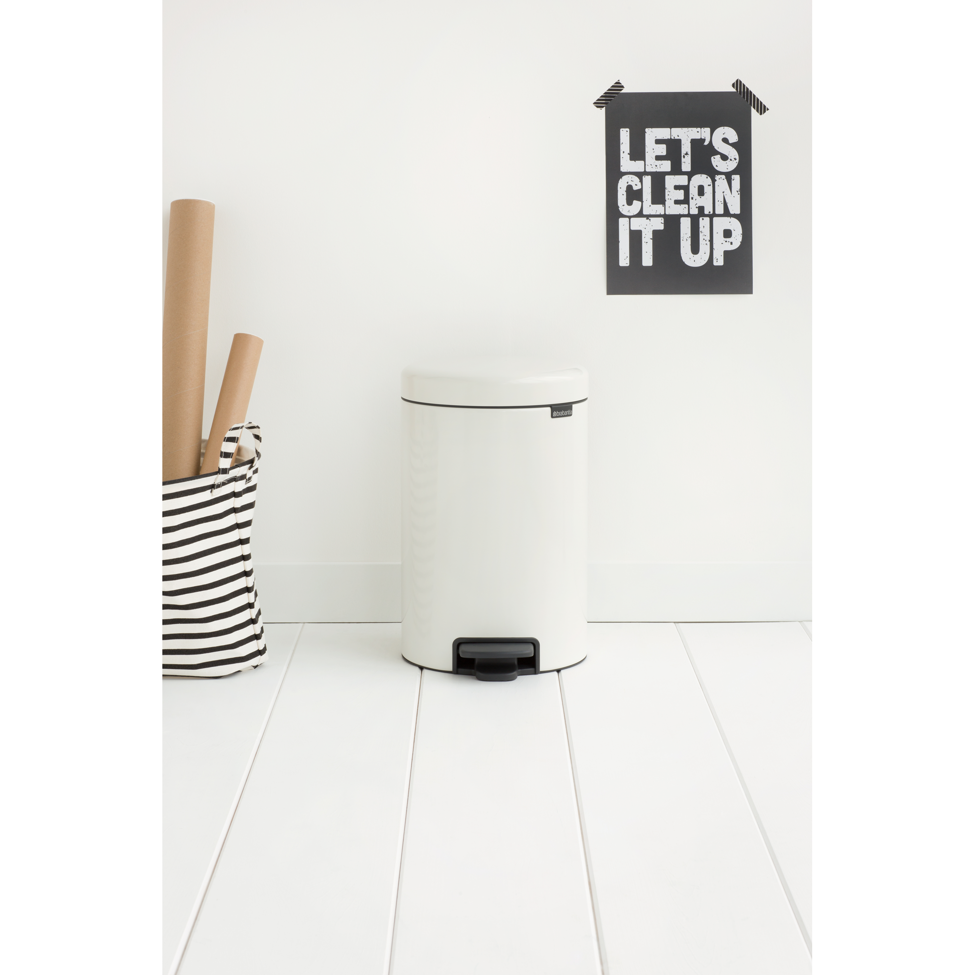 Een witte pedaalemmer staat op een witte houten vloer naast een gestreepte mand met kartonnen kokers.