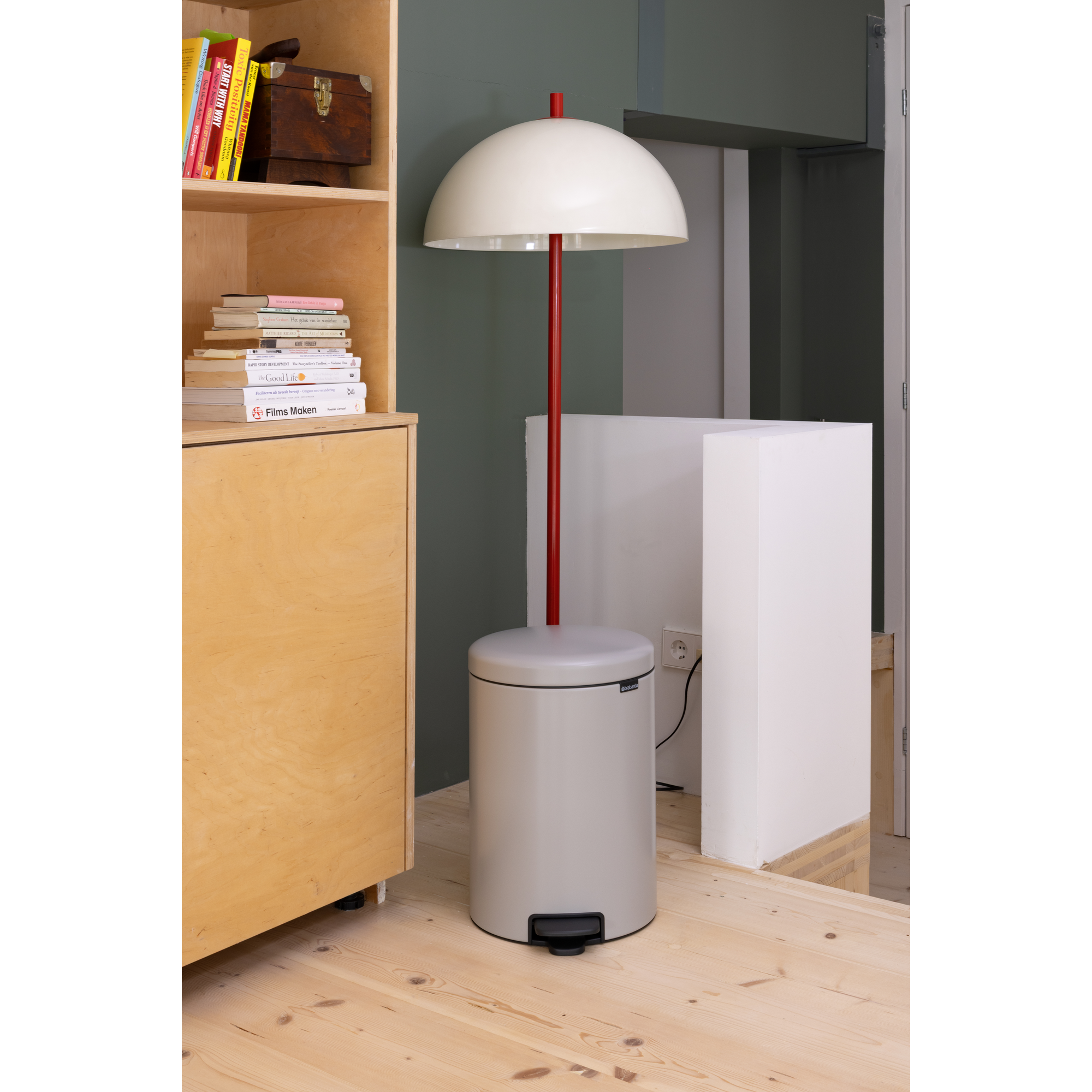 Een lichtgrijze pedaalemmer staat naast een boekenrek en een lamp.