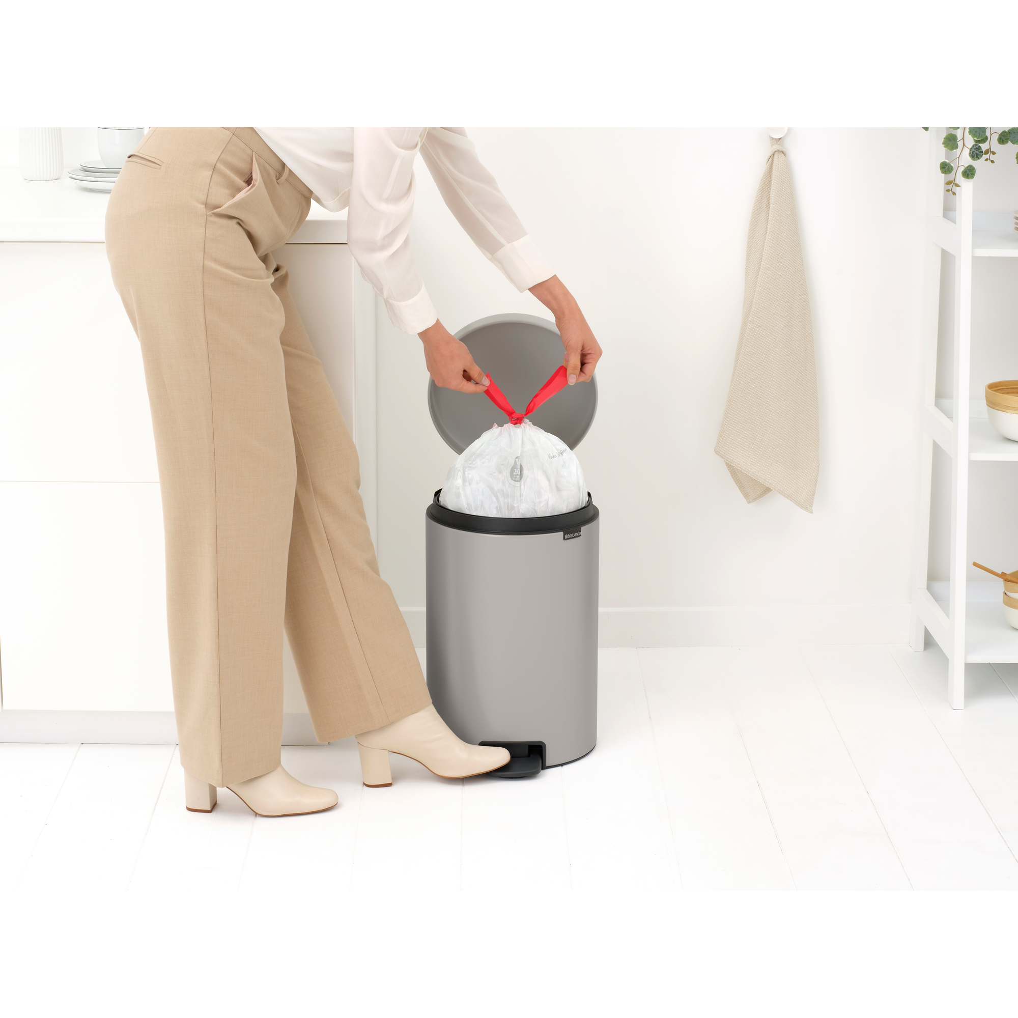 Een vrouw in een beige broek en witte schoenen knoopt een witte vuilniszak dicht in een prullenbak.