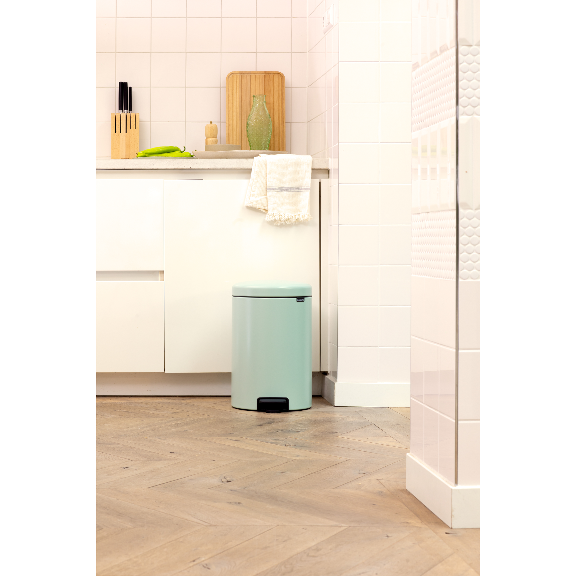 Un cubo de basura verde menta sobre suelo de madera frente a armarios de cocina blancos.