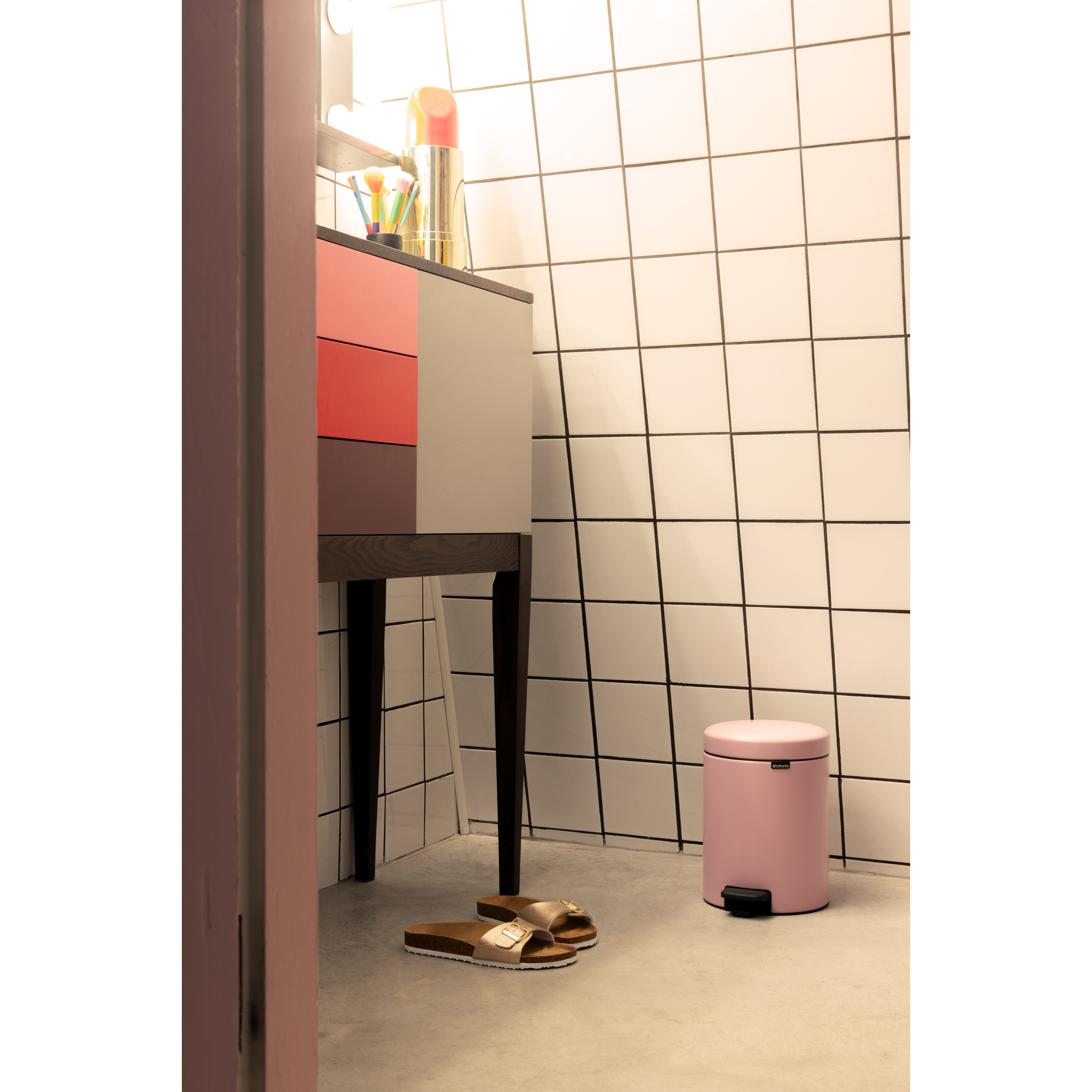 Een roze prullenbak naast een kast met make-upkwasten en een make-updispenser.