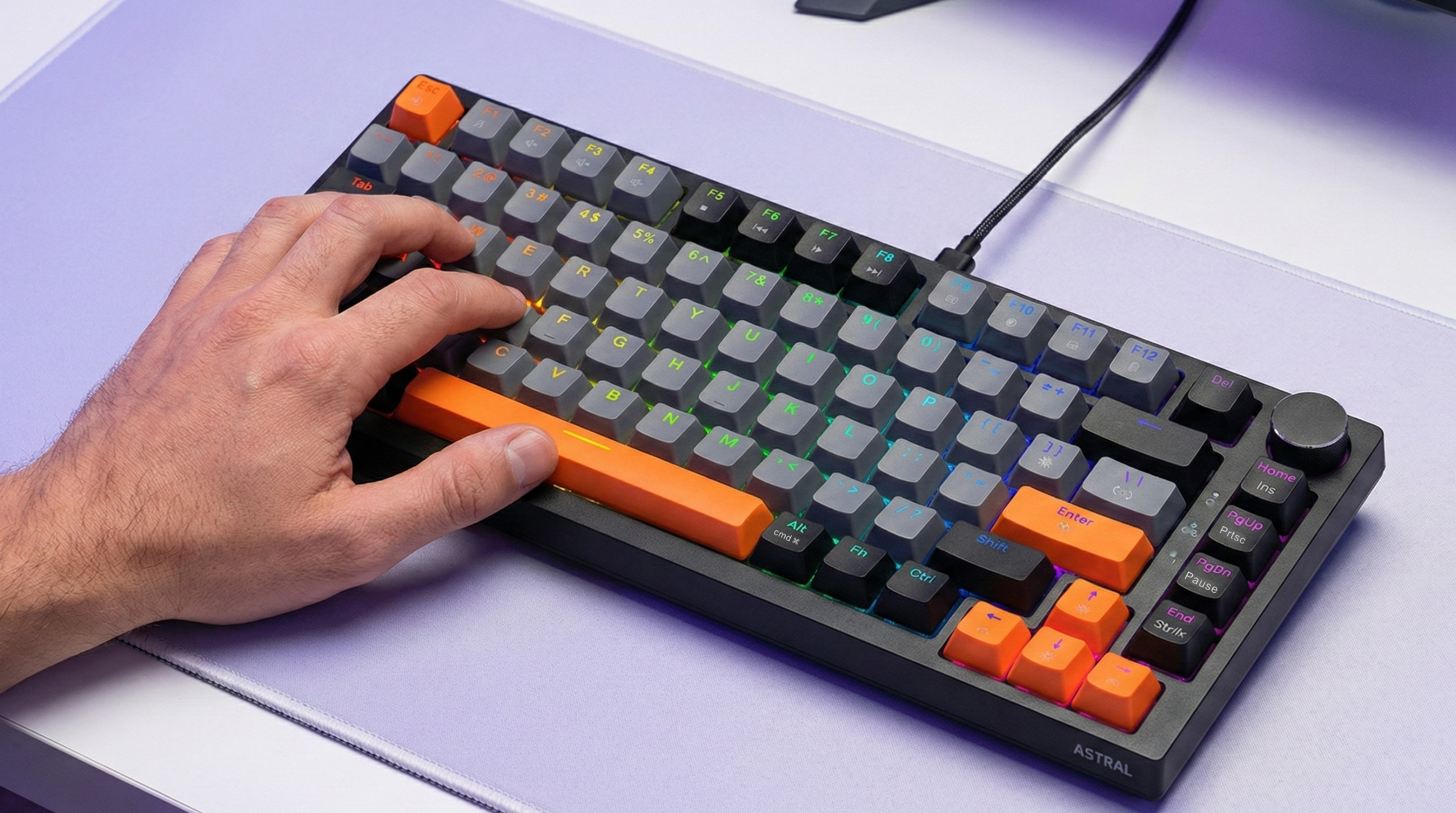 Dłoń spoczywa na czarnej klawiaturze mechanicznej z szarymi i pomarańczowymi klawiszami. Widoczne jest podświetlenie RGB.