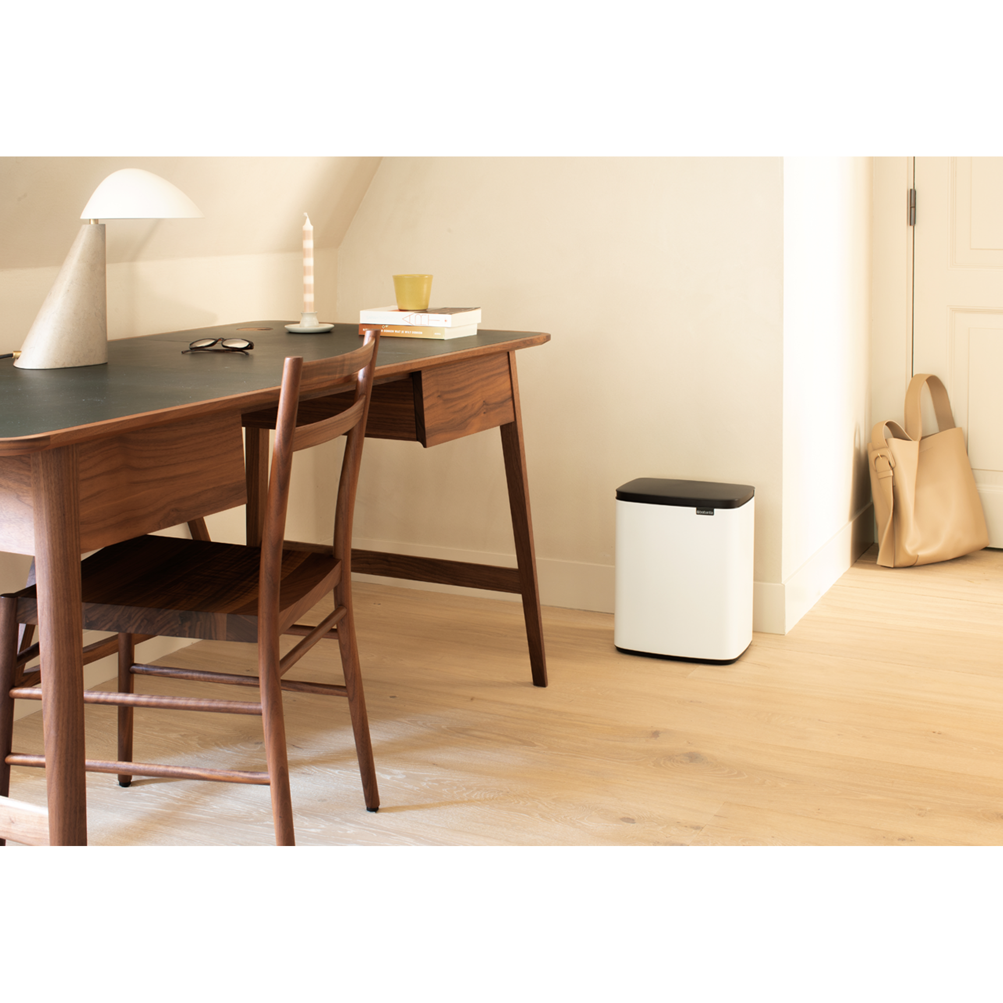 Een houten bureau met stoel, lamp, kaars, boeken, prullenbak en tas.