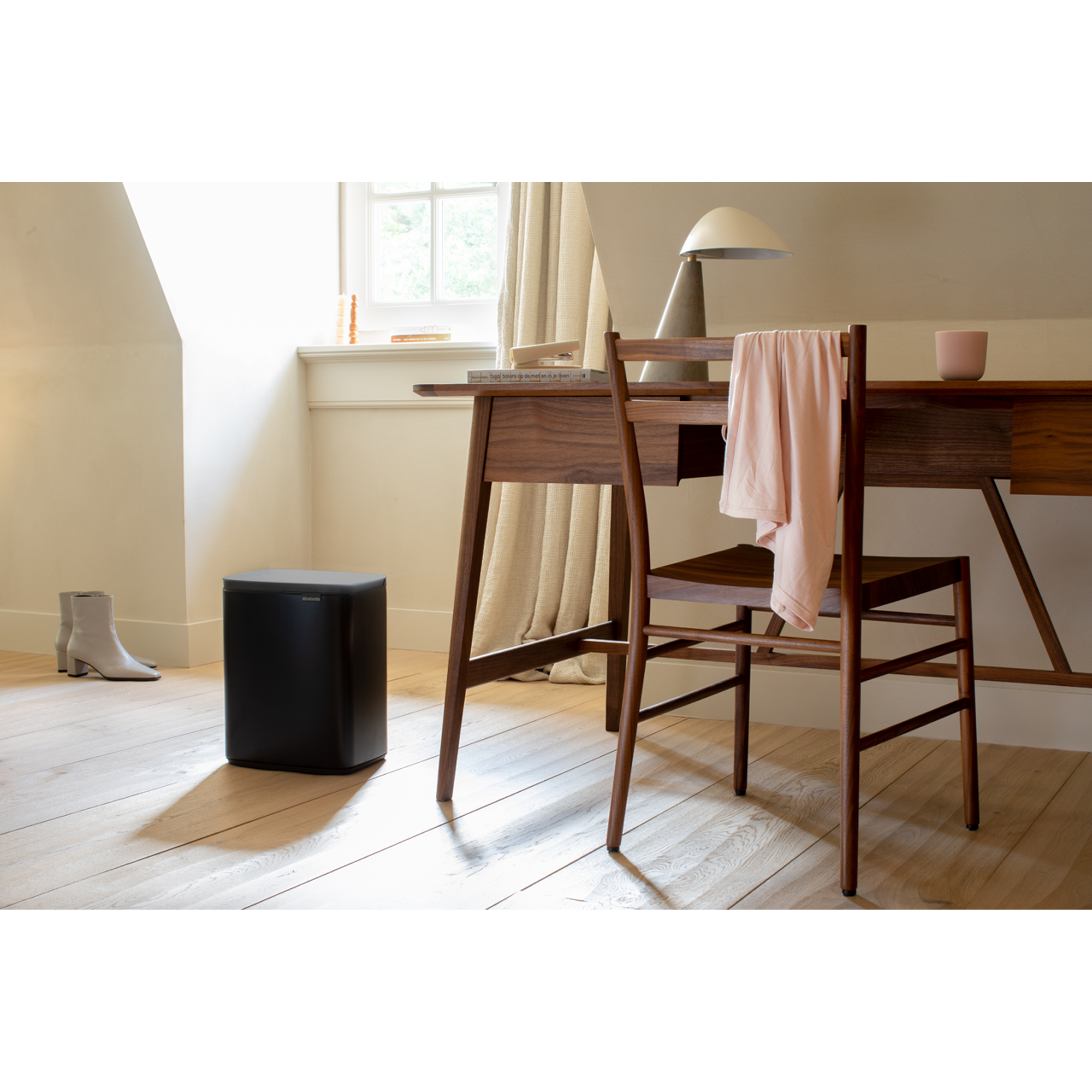 Een zwarte prullenbak en lichtbeige laarzen staan op een houten vloer. Een houten bureau met een stoel is in de buurt.