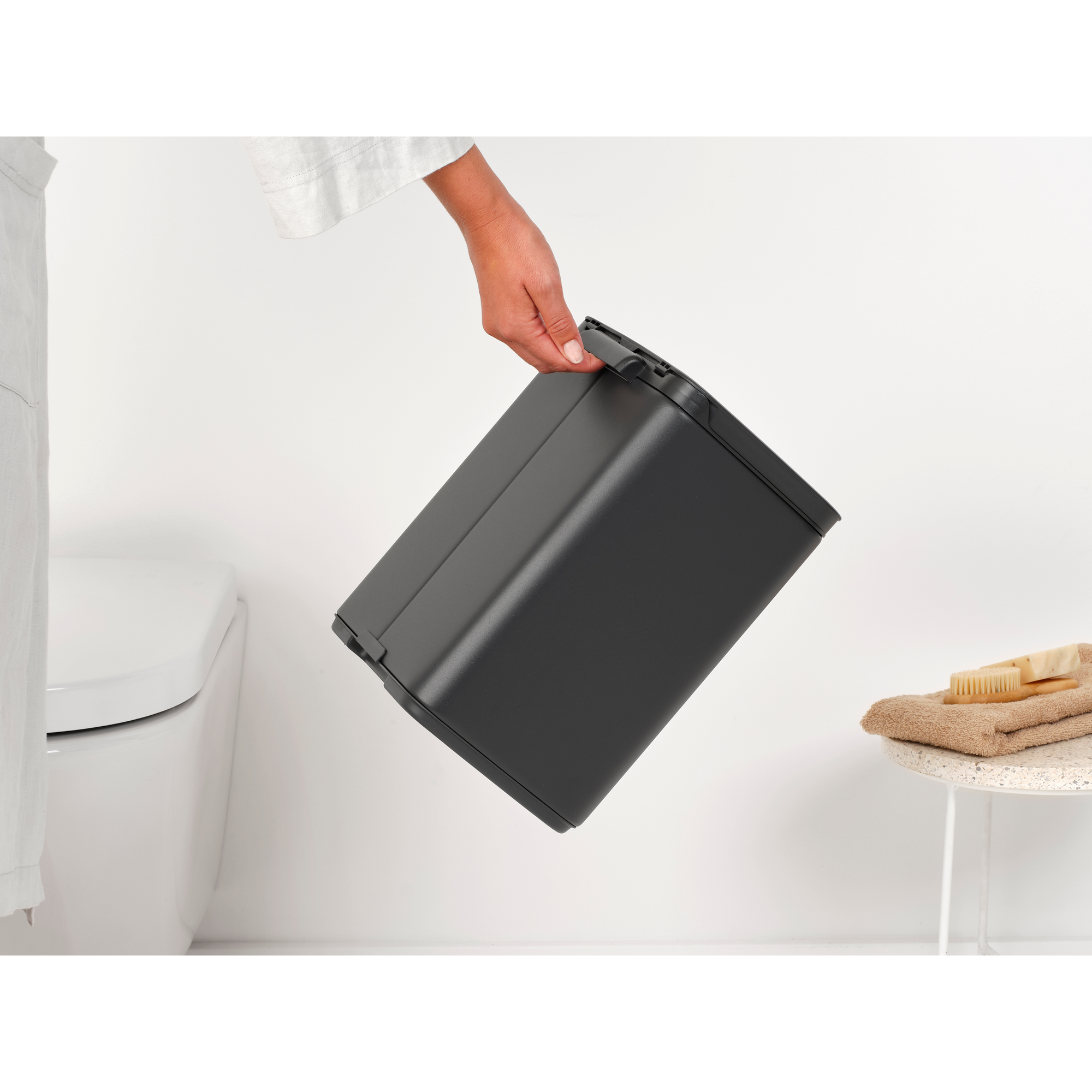 Een hand houdt een donkere prullenbak vast naast een wit toilet.