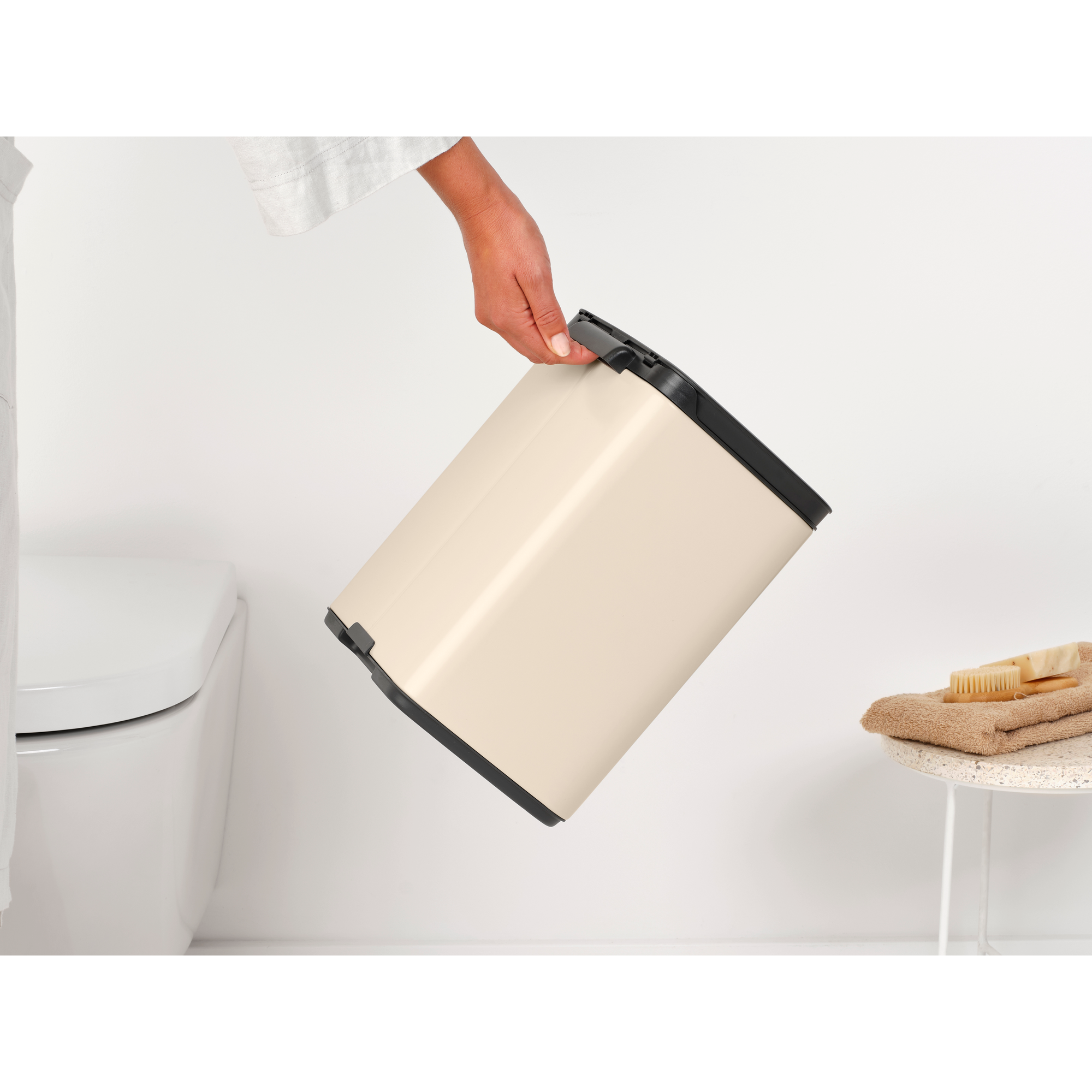 Een hand houdt een crème-kleurige prullenbak boven een toilet.
