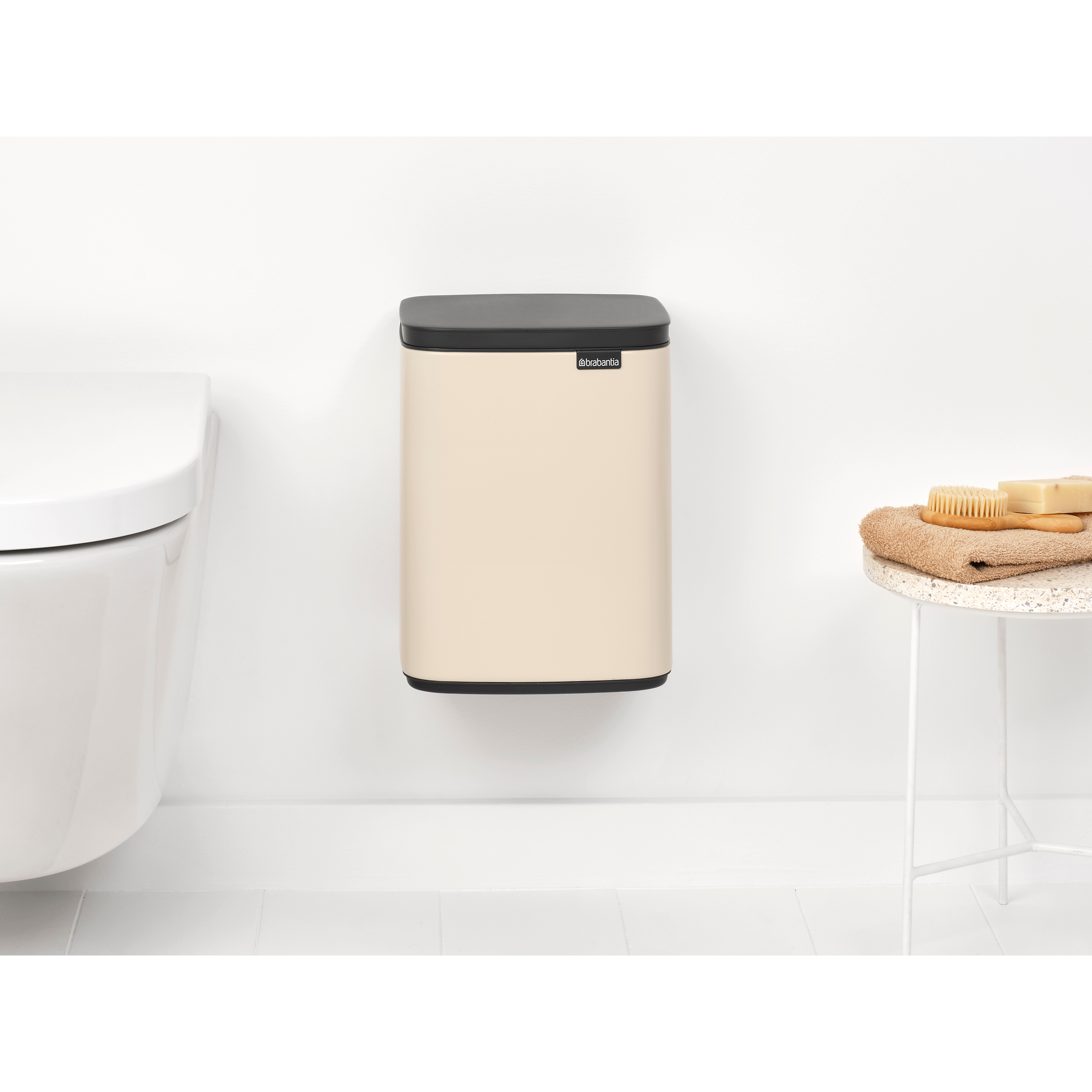 Een beige Brabantia prullenbak hangt aan een witte muur naast een toilet.