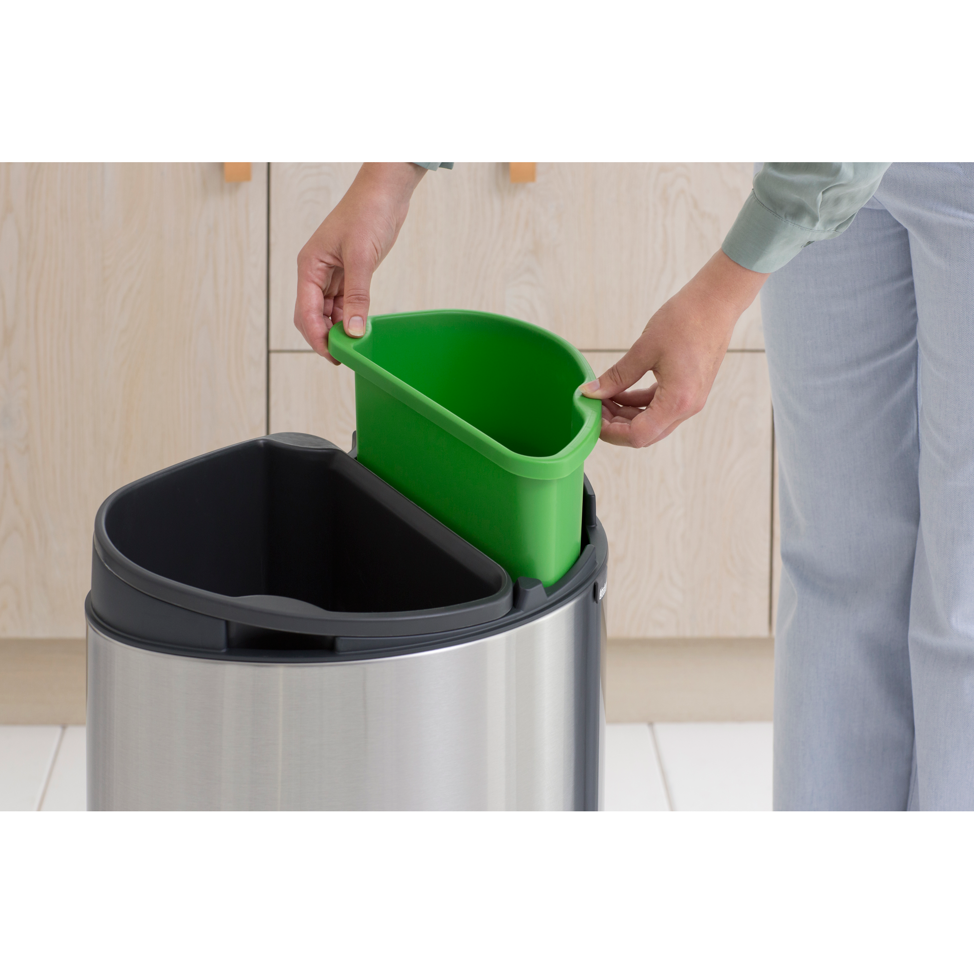 Een persoon plaatst een groene bak in een zilveren prullenbak.
