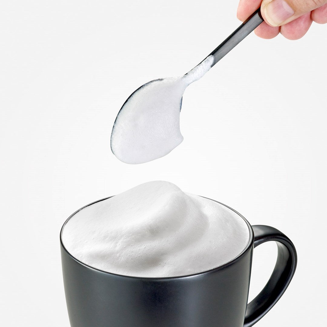 A spoon of milk foam above a black cup filled with milk foam.
