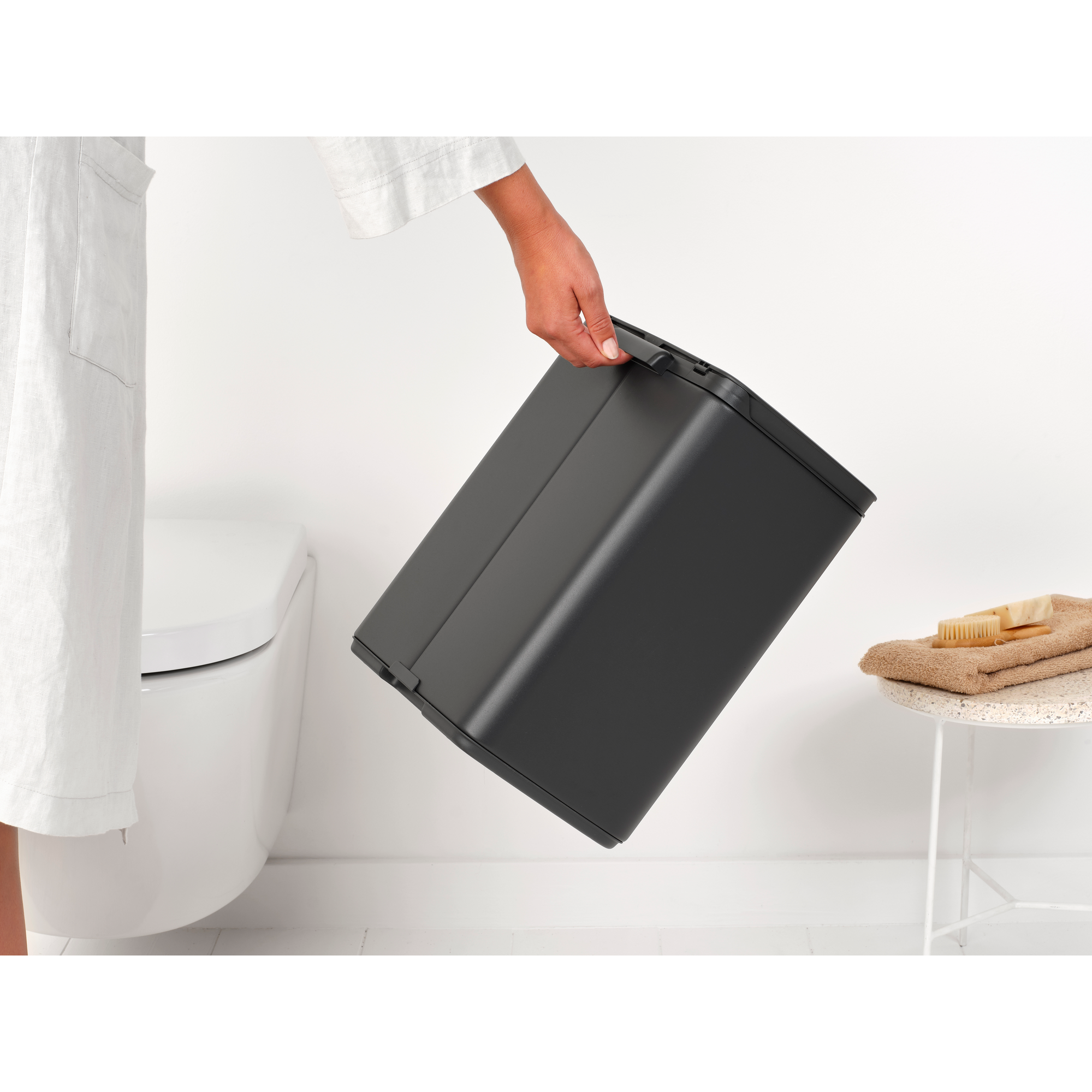 Een persoon houdt een donkere prullenbak vast naast een toilet in een badkamer.