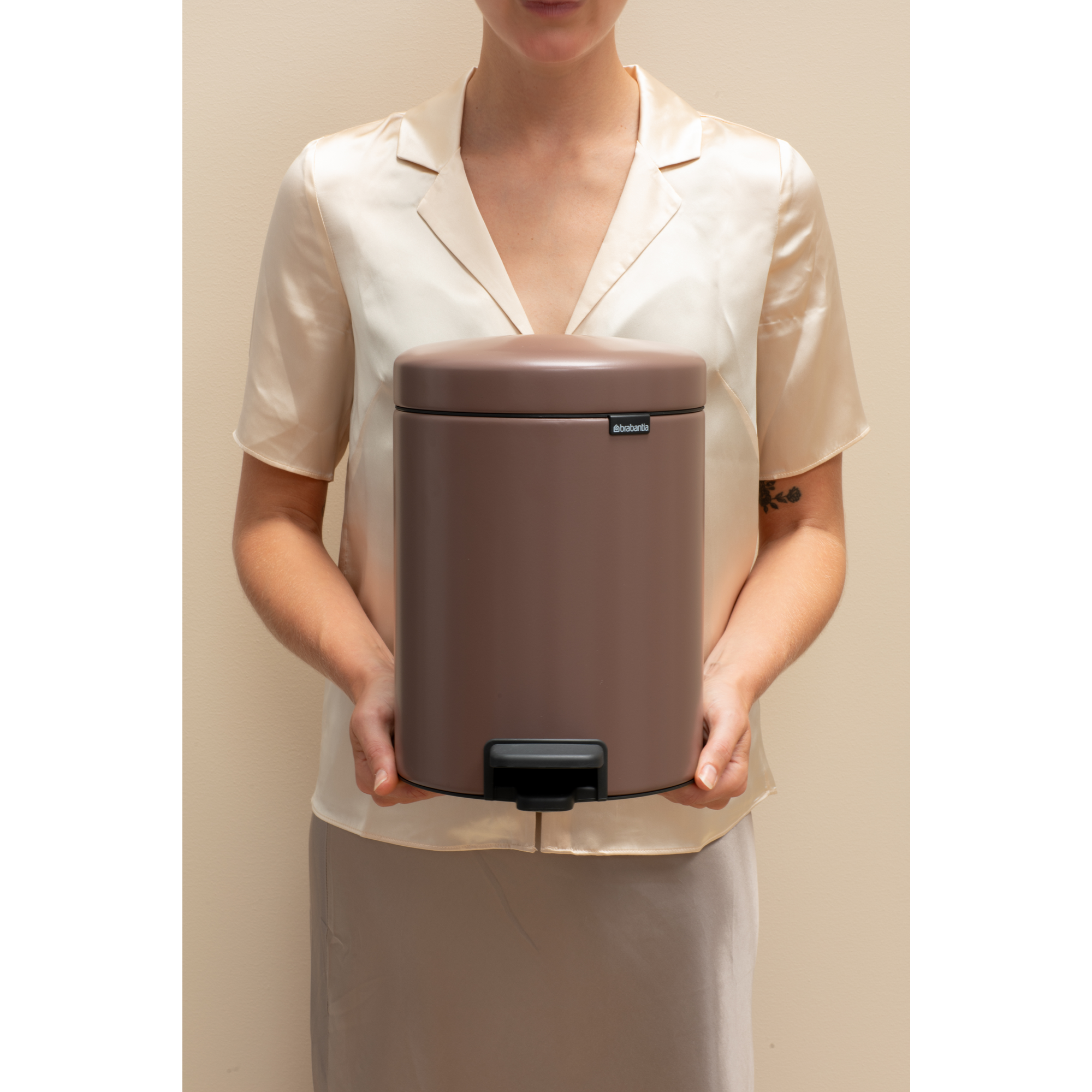 Een vrouw houdt een bruine prullenbak met voetpedaal en zwart pedaal vast tegen beige achtergrond.