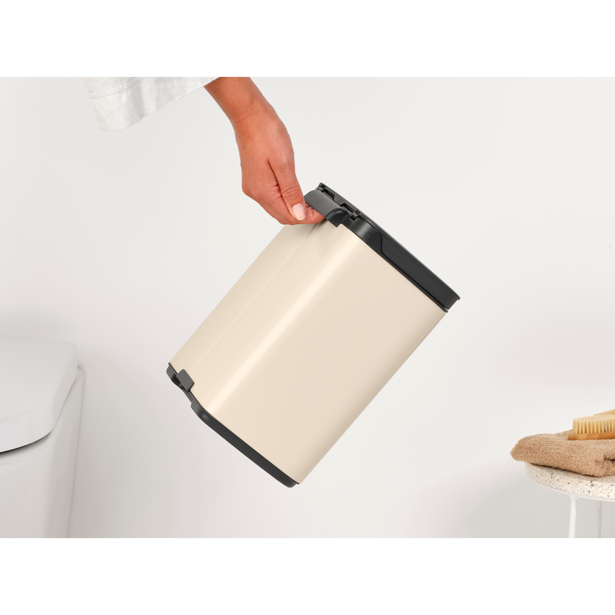 Een hand houdt een crèmekleurige prullenbak vast naast een toilet.