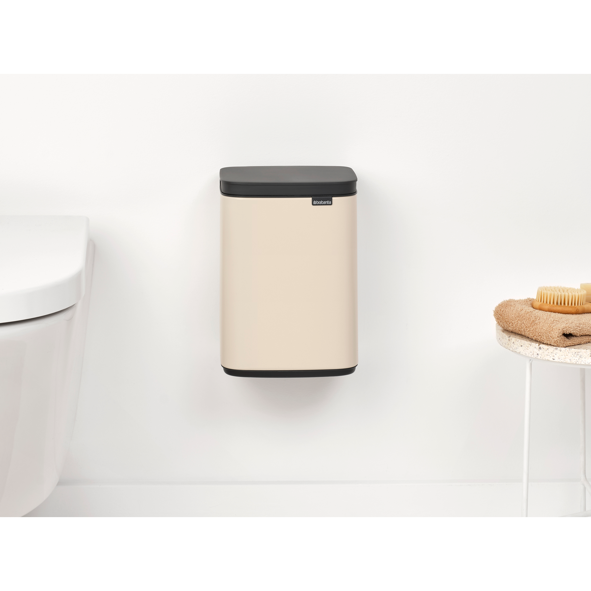 Beige wandprullenbak met zwart deksel, naast een toilet.