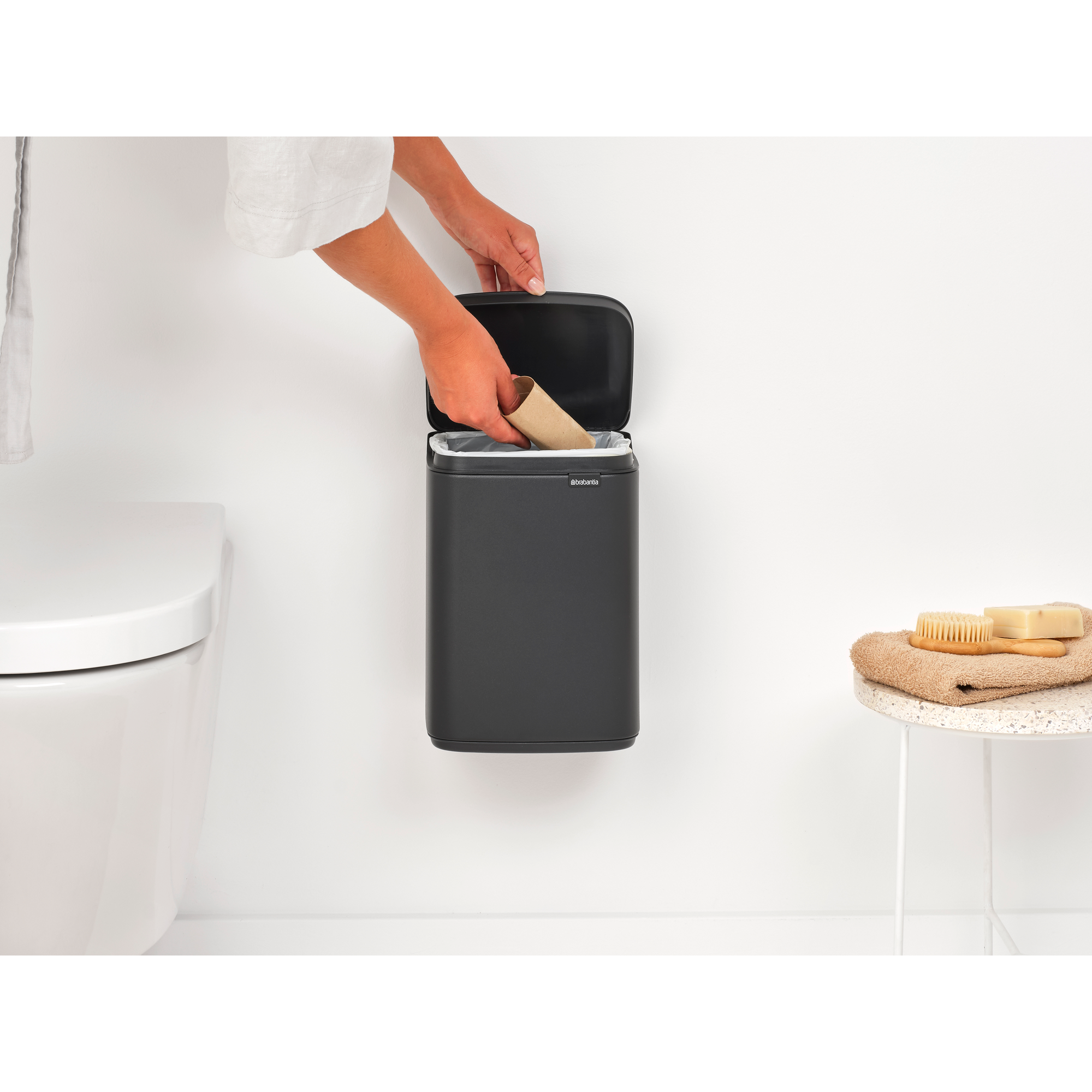 Een hand gooit een lege wc-rol in een donkere prullenbak naast een toilet.