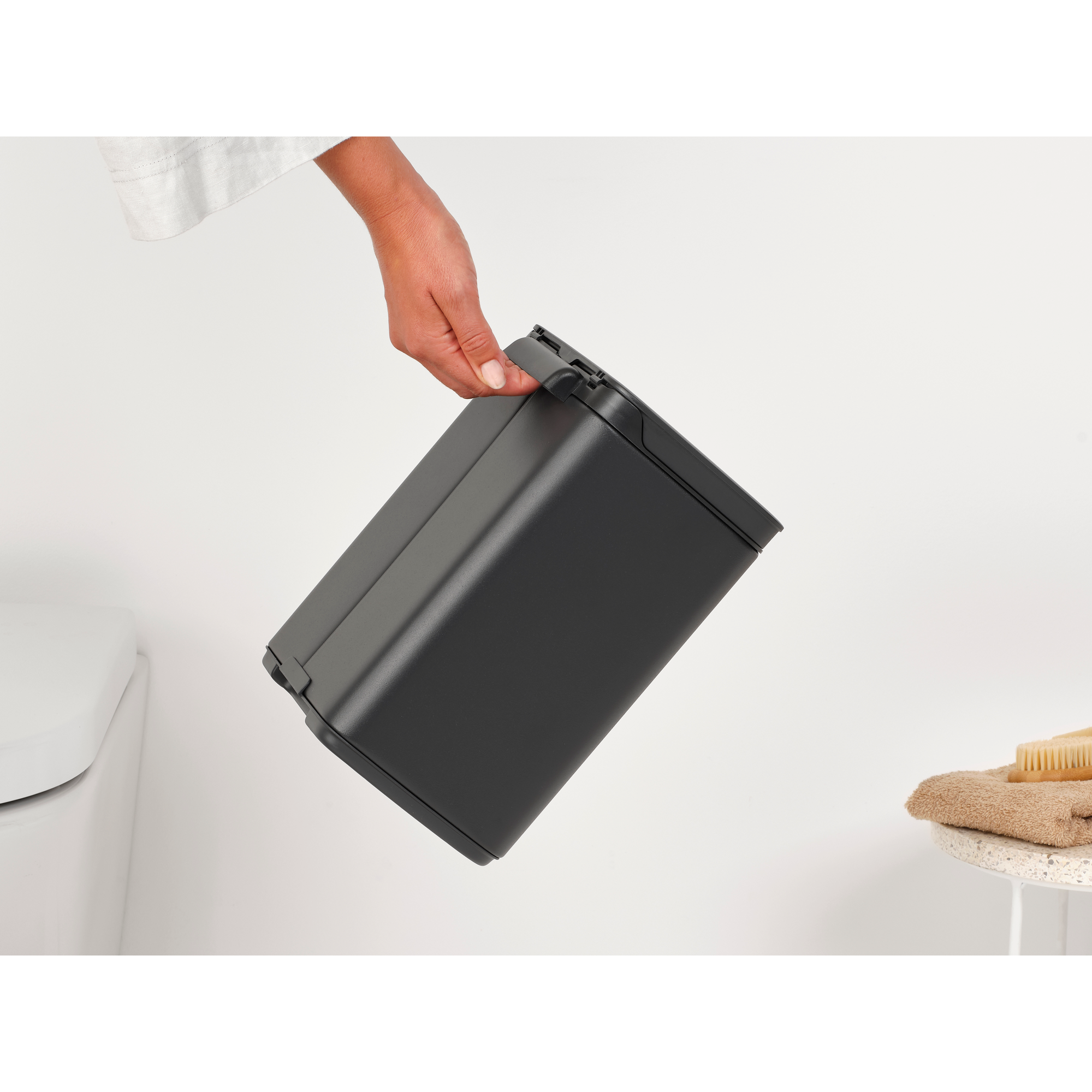 Een hand tilt een zwarte prullenbak op naast een toilet. Een handdoek en borstel zijn zichtbaar.