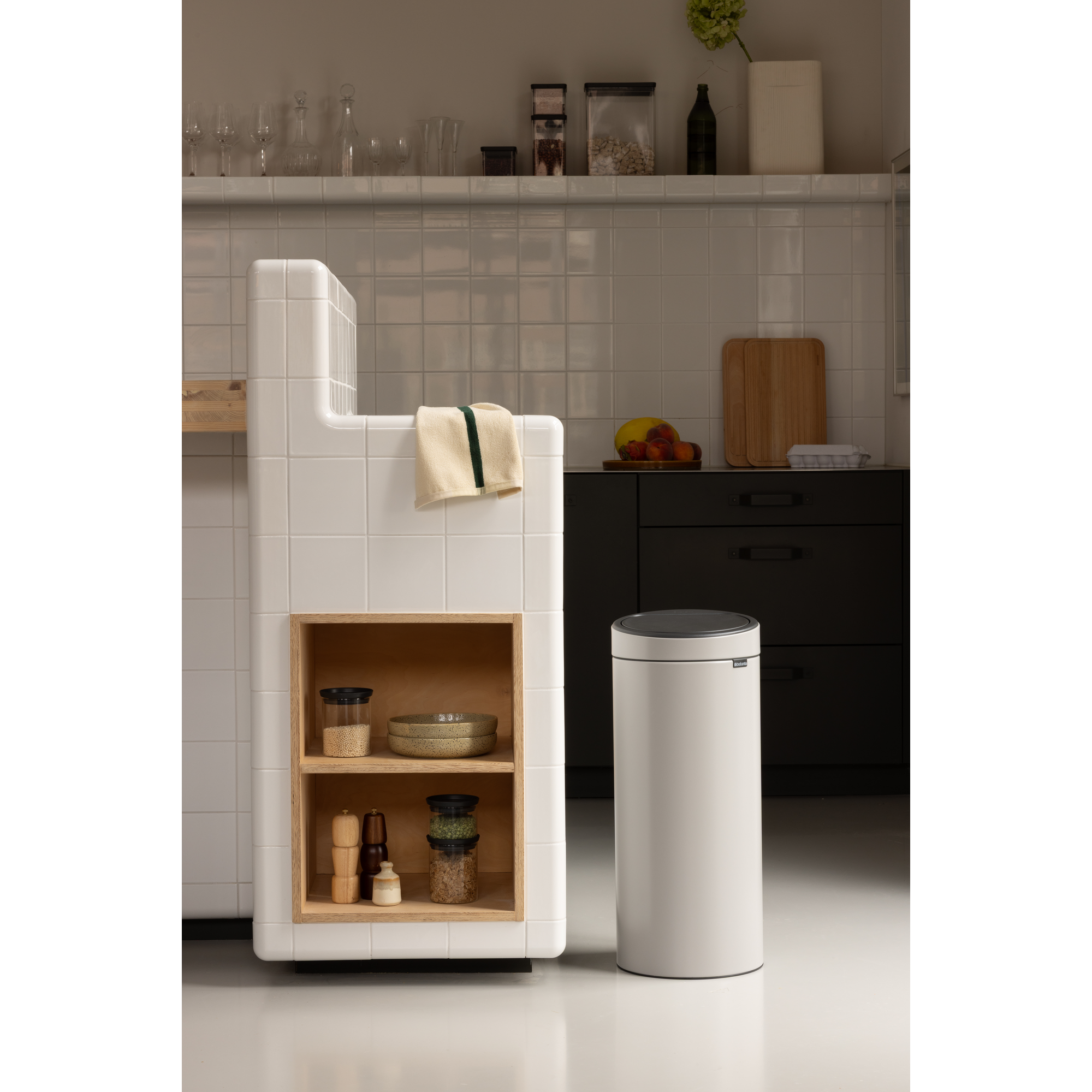 Moderne keuken met witte prullenbak, houten plank met voorraad en witte tegels.