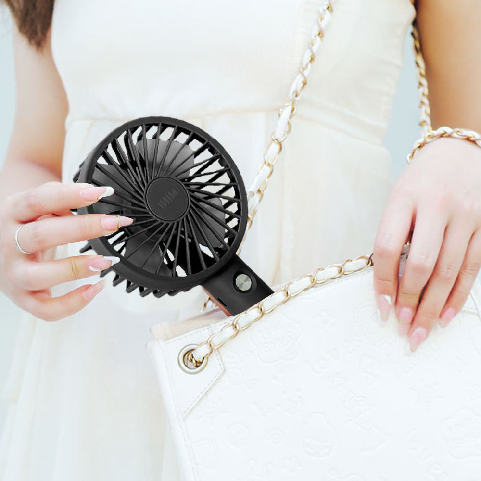Eine Person im weißen Kleid hält einen schwarzen Ventilator in der Nähe einer weißen Tasche mit goldener Kette.