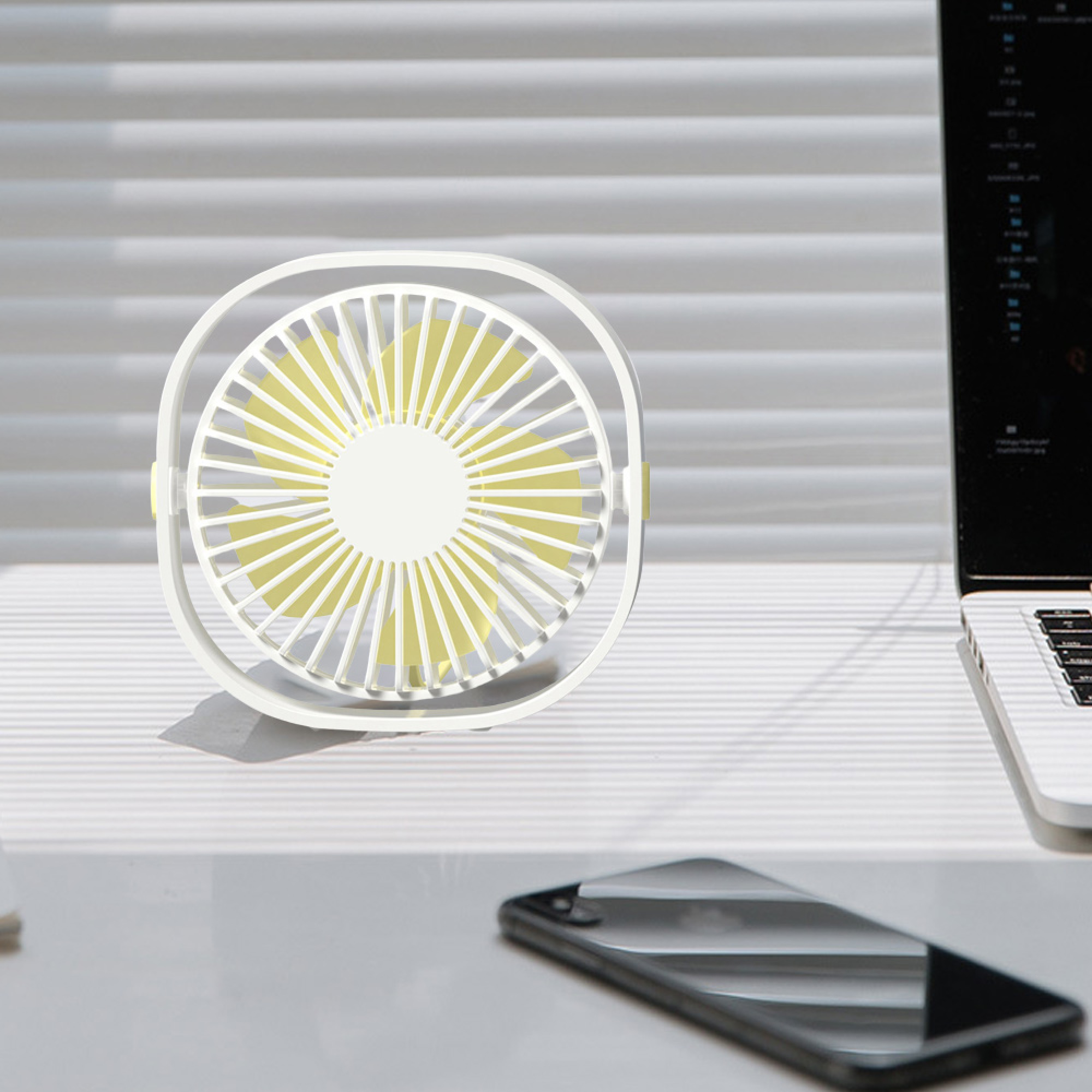 Ein weißer Tischventilator mit gelben Rotorblättern steht auf einem Schreibtisch neben einem Laptop und einem Smartphone.