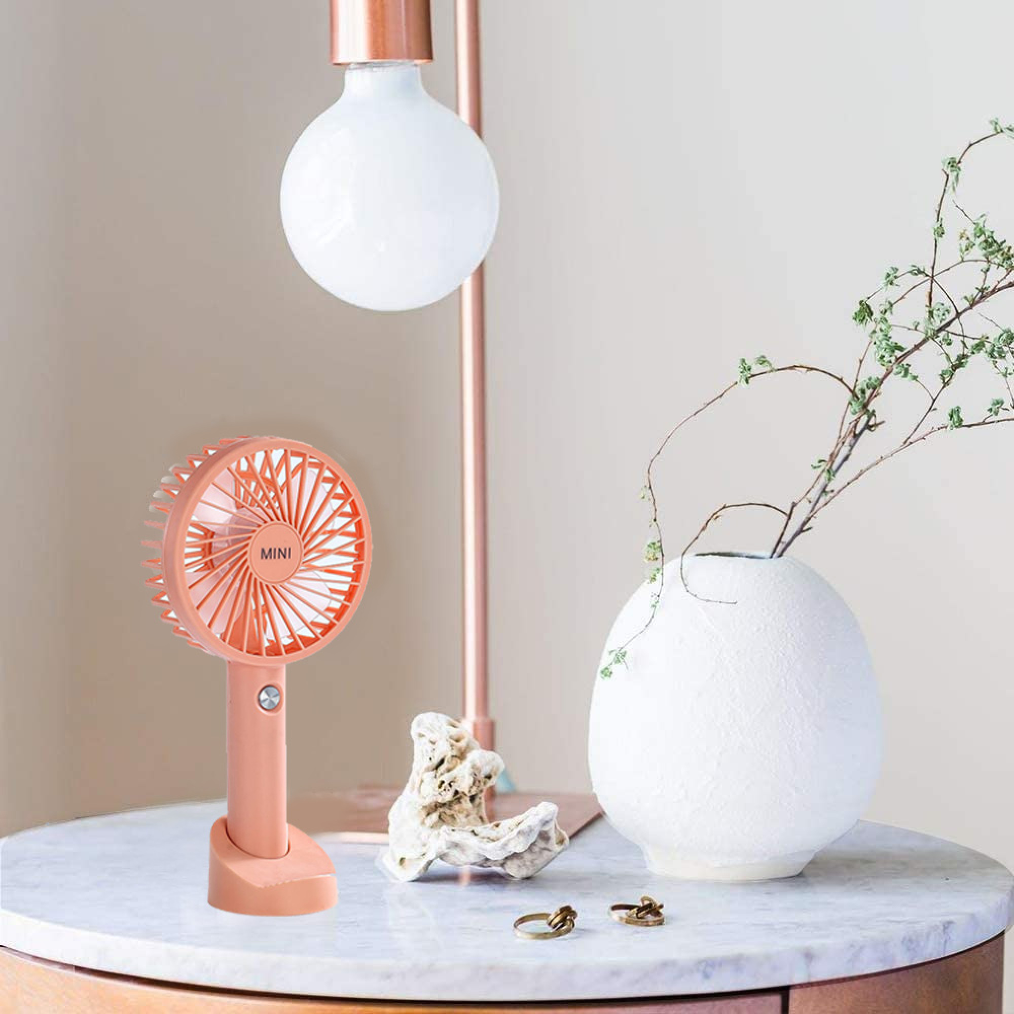 Ein kleiner rosa Ventilator steht auf einem Marmortisch neben einer Vase mit Zweigen.