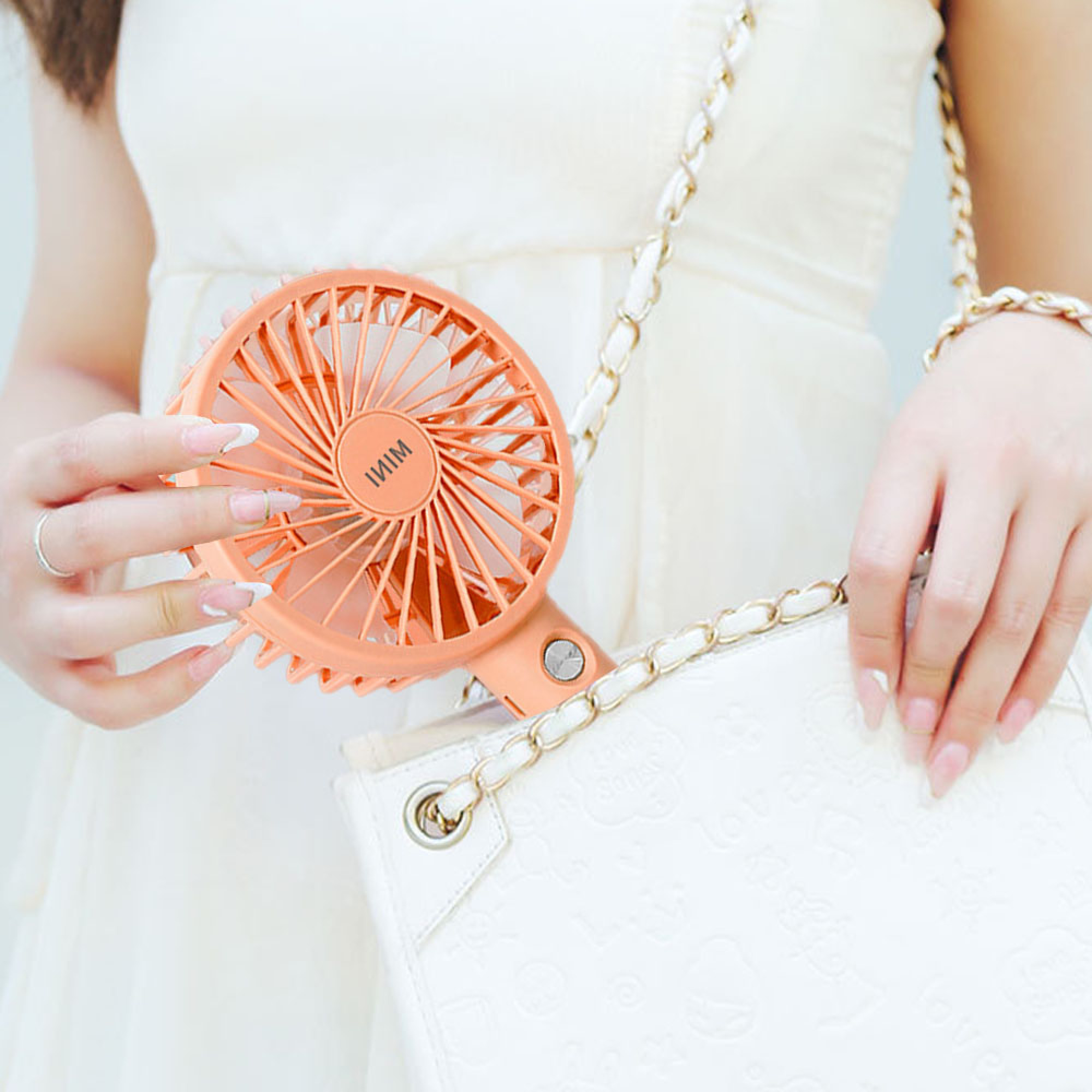 Eine Person hält einen orangefarbenen Mini-Ventilator neben einer weißen Handtasche mit Kettenriemen.