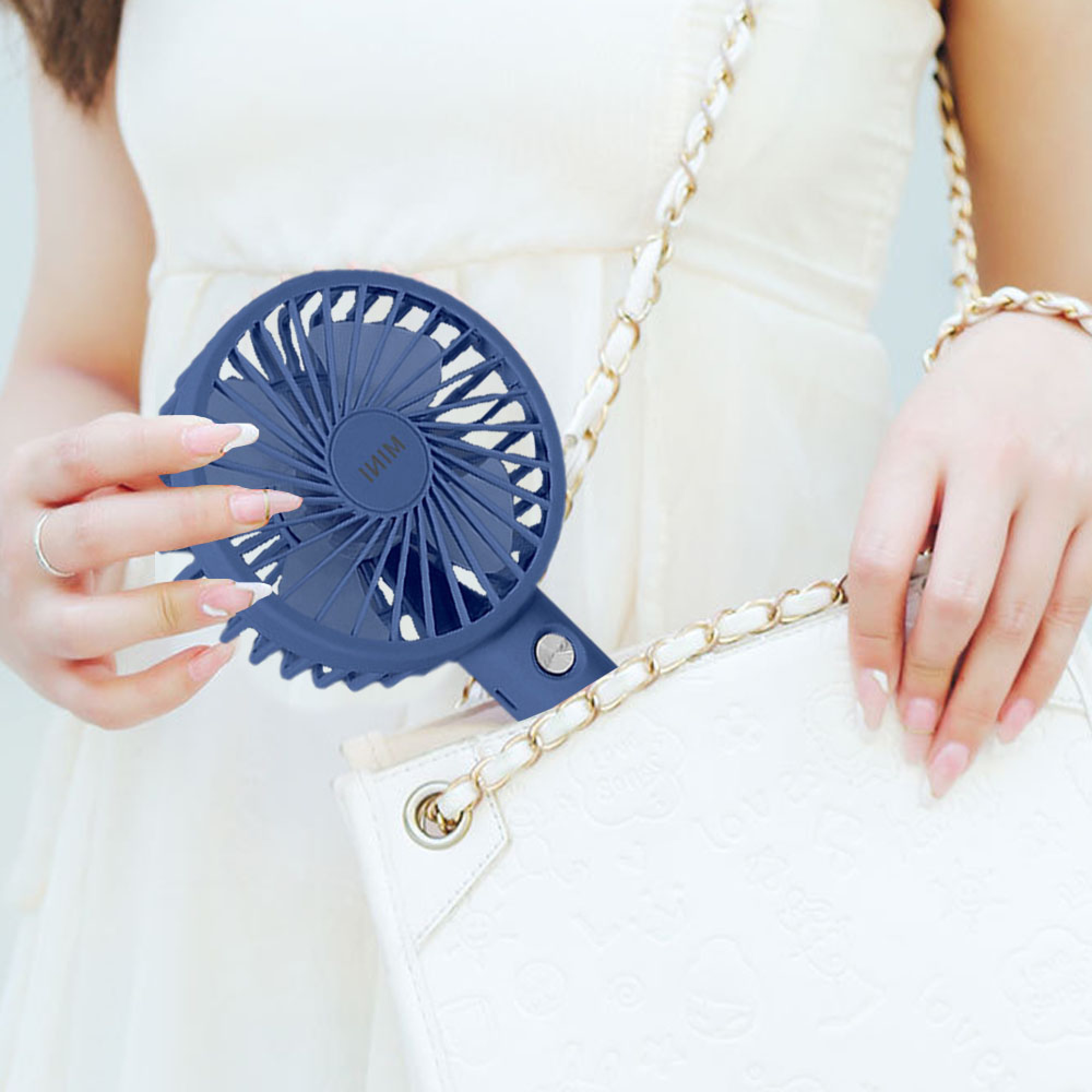 Eine Frau hält einen blauen Handventilator und eine weiße Tasche mit goldener Kette.