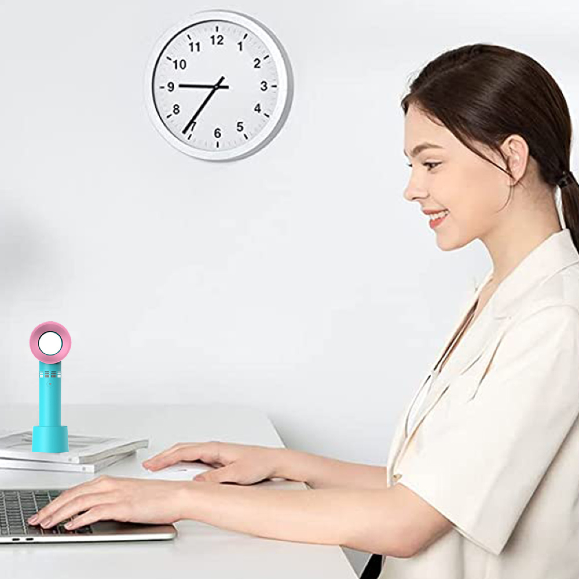 Eine Frau tippt auf einem Laptop, daneben ein kleiner Ventilator und eine Wanduhr.