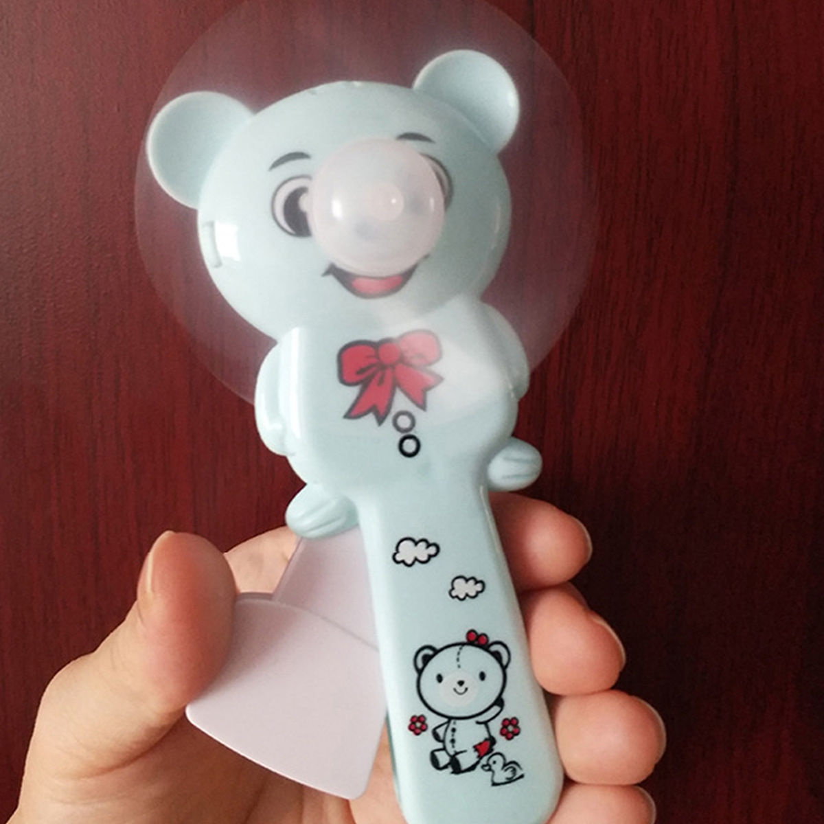 Ein türkisfarbener Teddybär-Ventilator mit einem Ball im Mund, der von einer Hand gehalten wird.