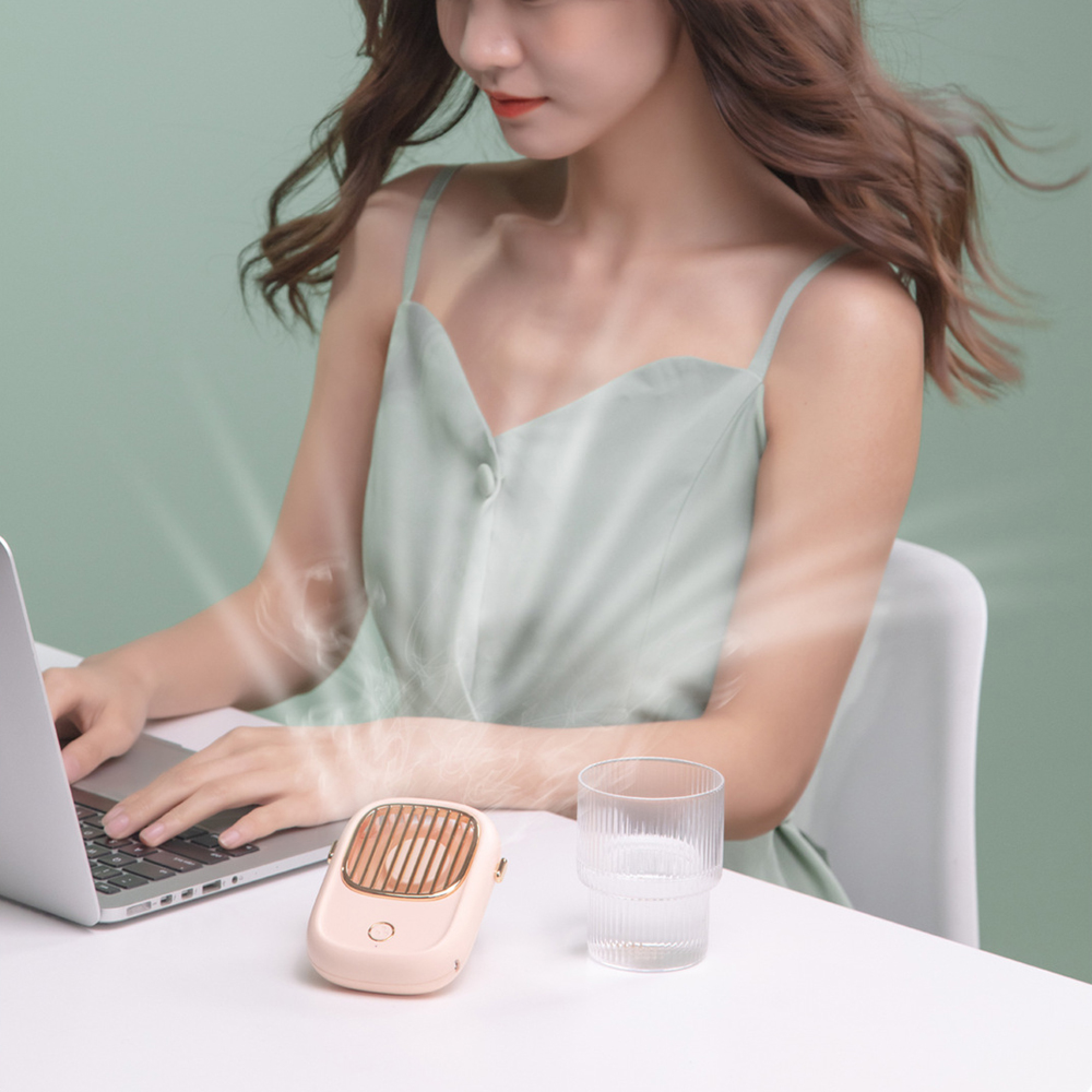 Eine Frau tippt auf einem Laptop mit einem rosa Ventilator und einem Glas Wasser auf einem Schreibtisch.
