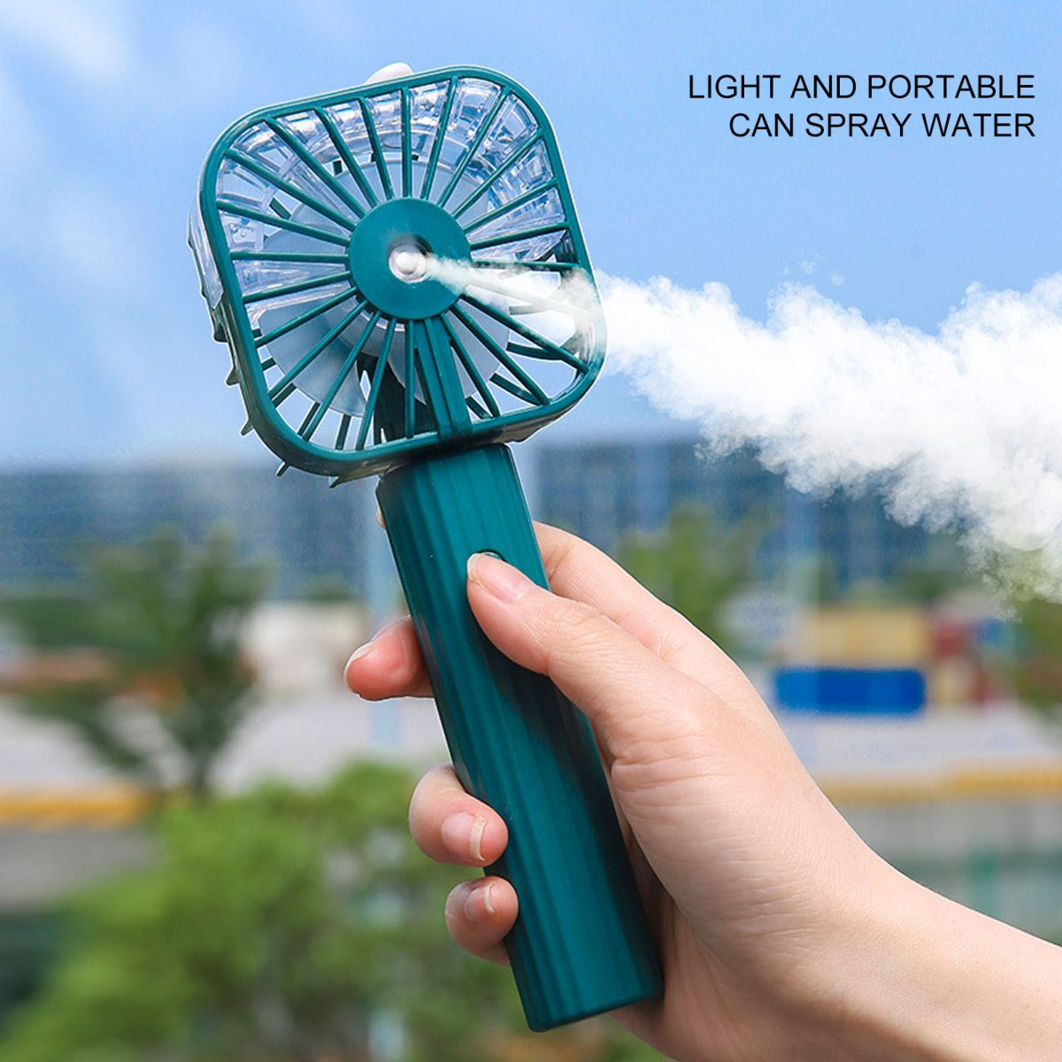 Eine Hand hält einen teal tragbaren Ventilator, der Nebel versprüht, vor einem unscharfen Hintergrund aus Gebäuden und Grün.