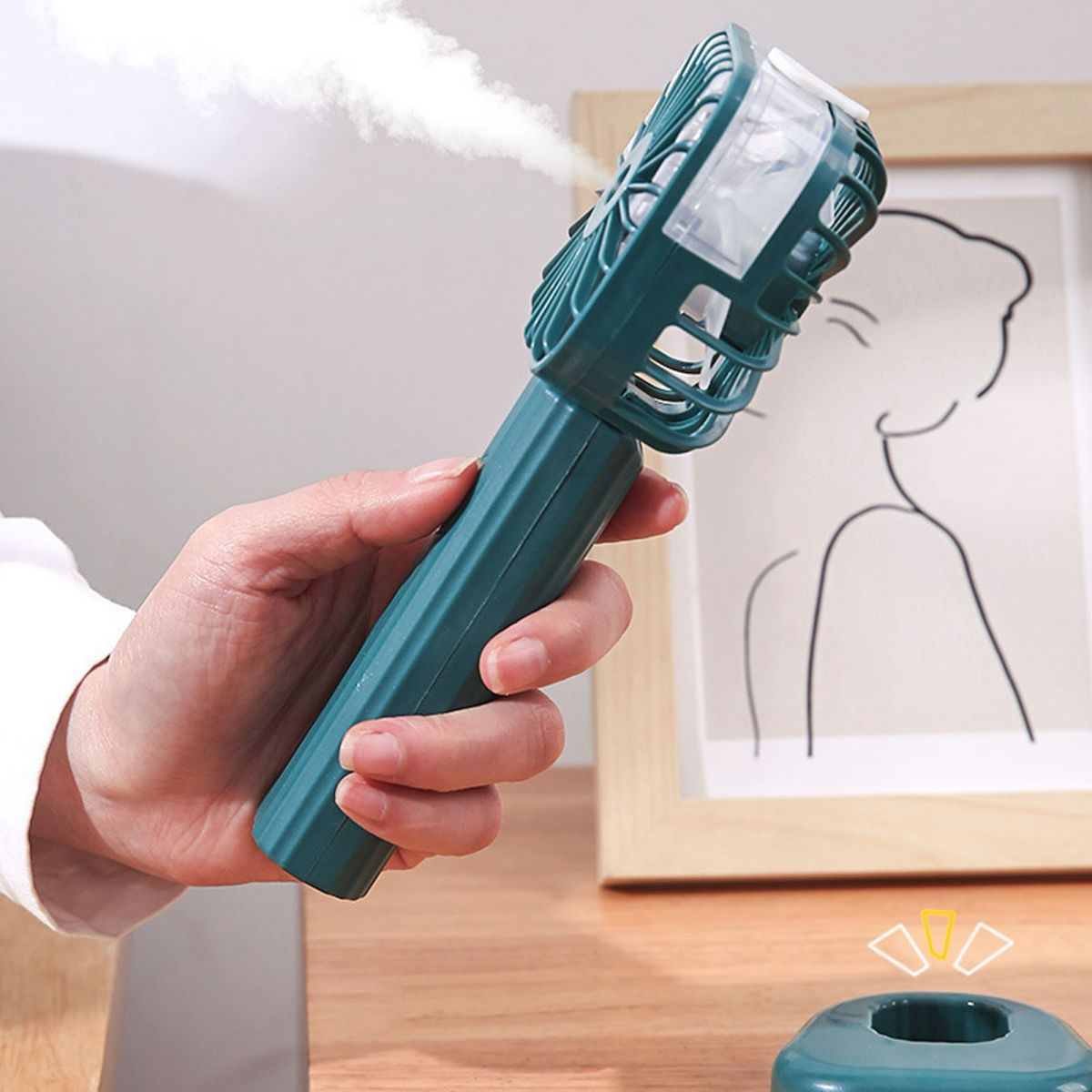 Eine Hand hält einen tealfarbenen Ventilator, aus dessen Oberseite Nebel sprüht. Ein minimalistischer Rahmen dahinter.