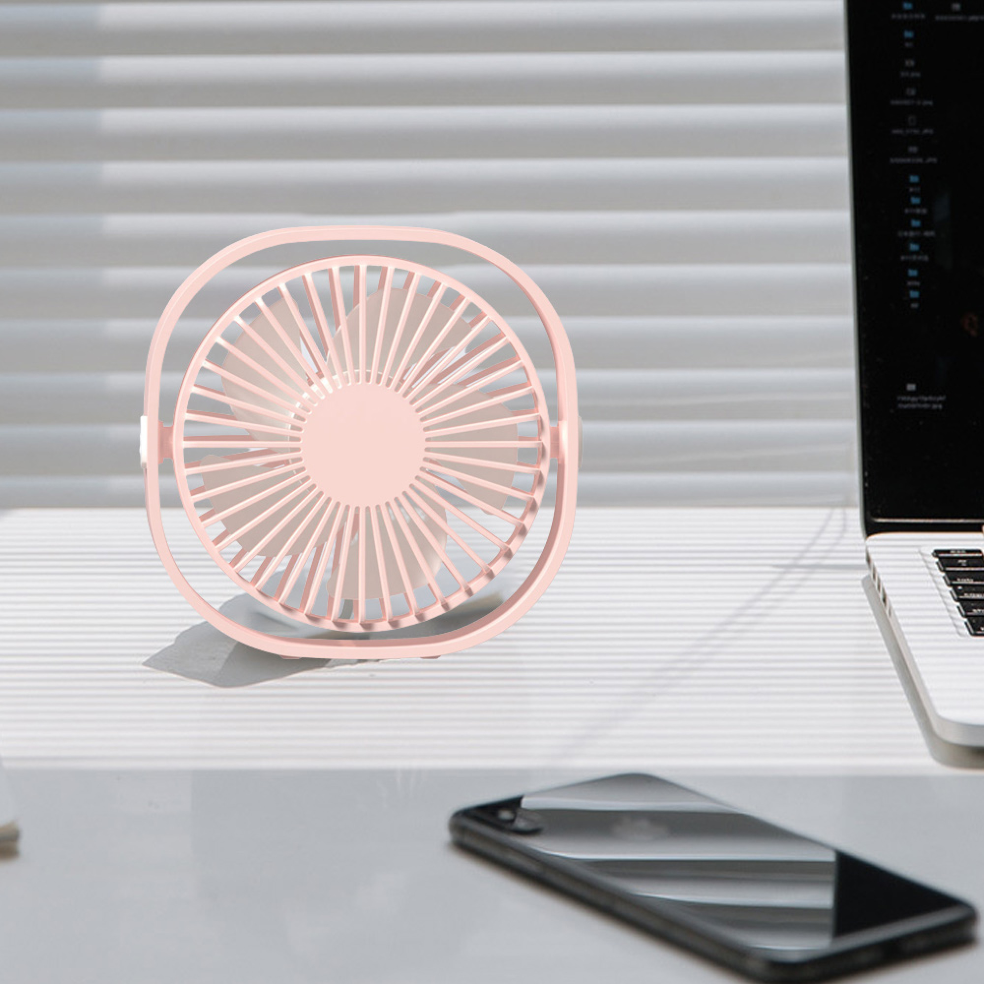 Ein kleiner rosa Ventilator steht auf einem Schreibtisch neben einem Laptop und Handy.
