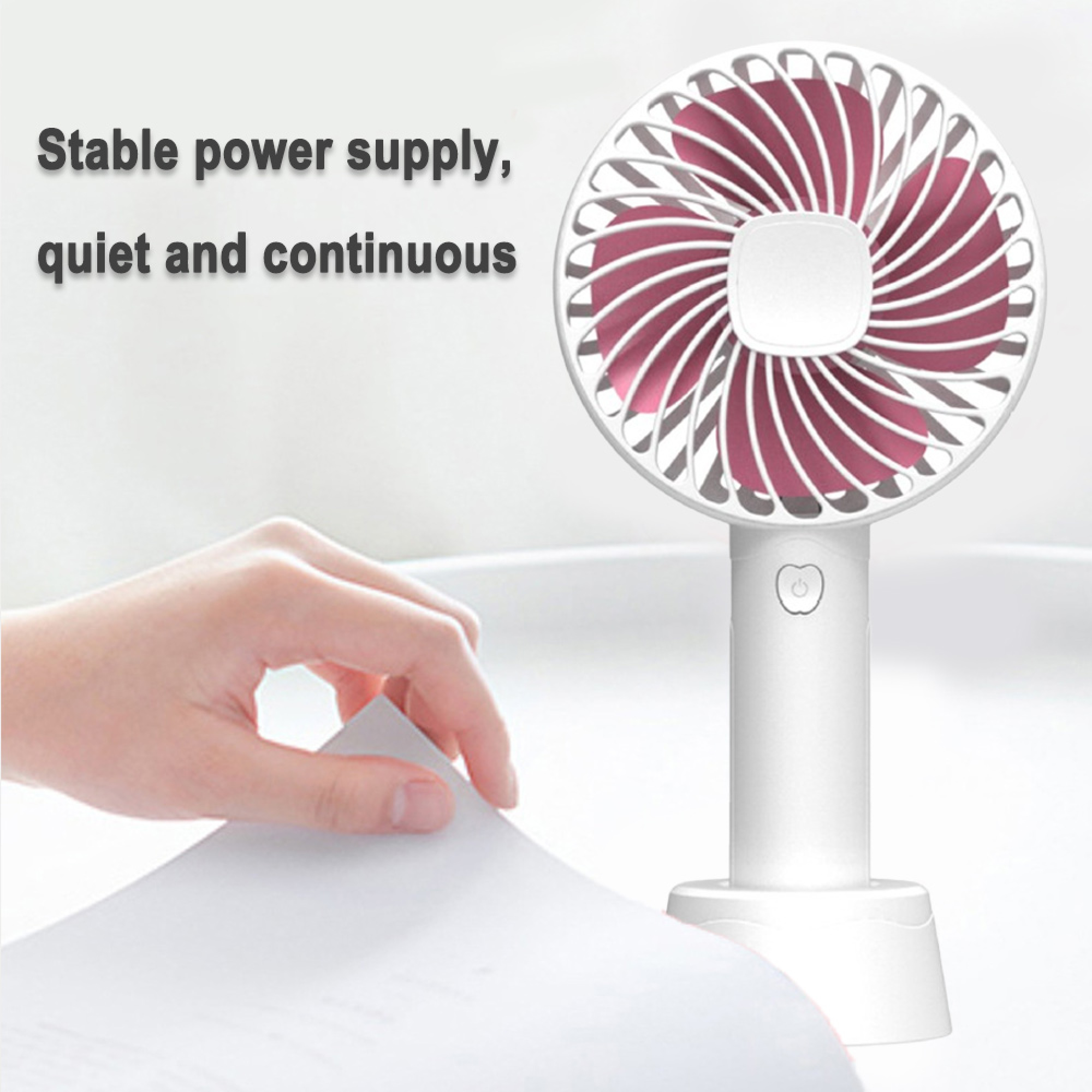 Ein weißer Handventilator mit rosa Rotorblättern steht auf einem Sockel. Eine Hand hält ein Blatt Papier.