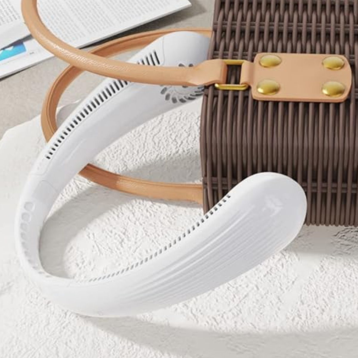 Ein weißer Nackenventilator liegt neben einer braunen gewebten Tasche auf einer texturierten Oberfläche.