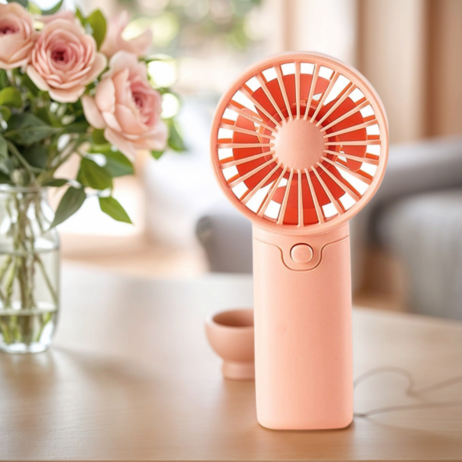 Ein rosafarbener Handventilator steht neben einer Vase mit rosa Rosen auf einem Holztisch.