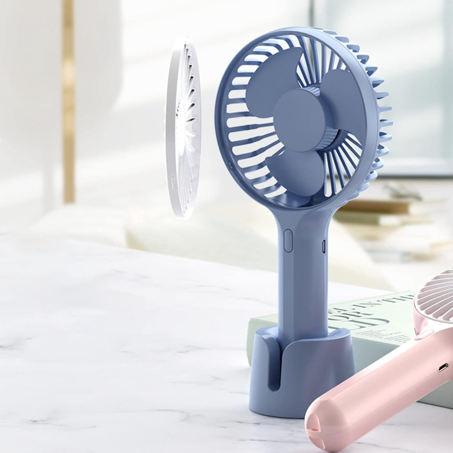Zwei tragbare Ventilatoren, blau und rosa, auf einem Schreibtisch.