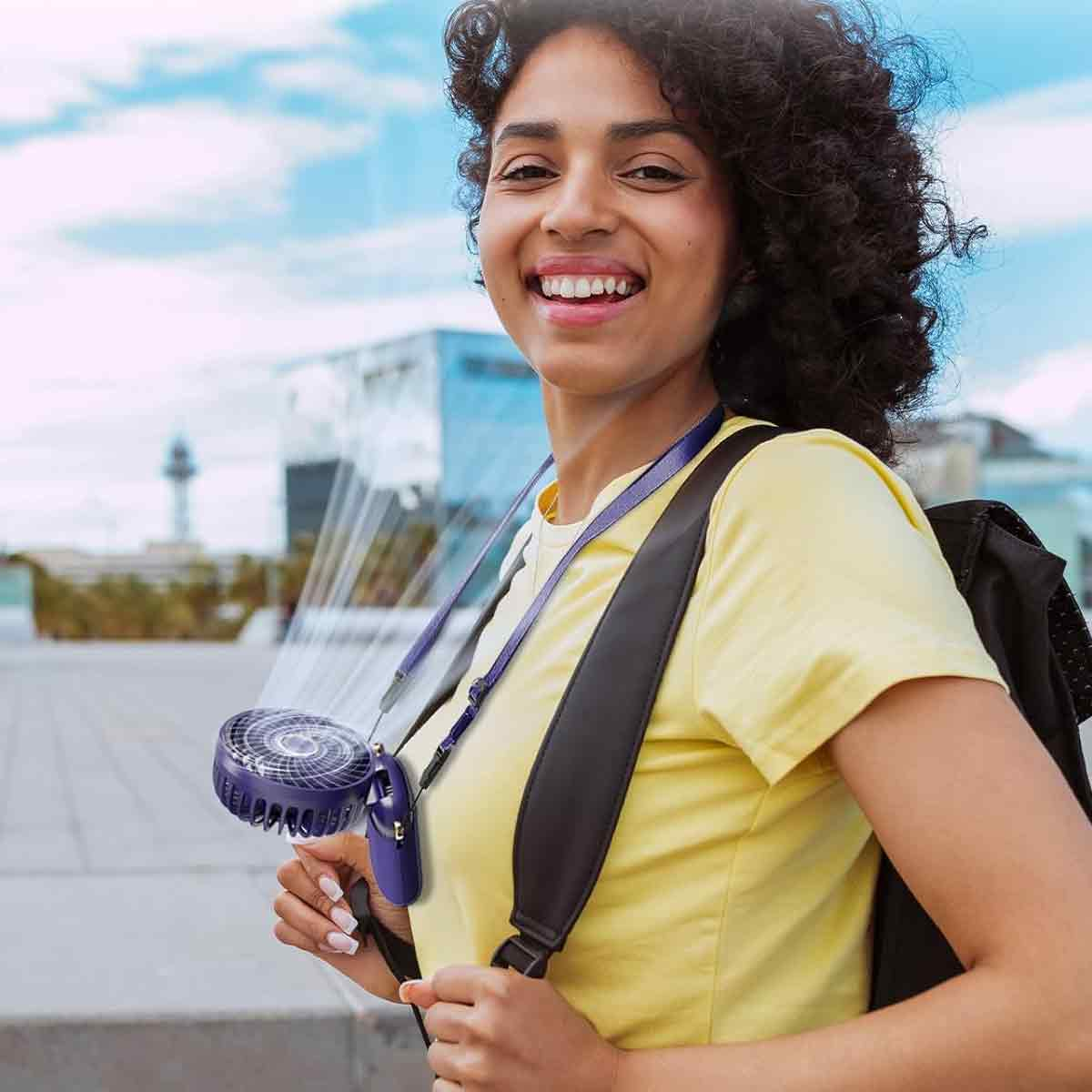 Frau mit Halsventilator, gelbes T-Shirt, Rucksack, Stadt im Hintergrund.